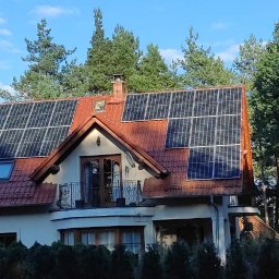 Sun Tech Fotowoltaika Rainczuk Sebastian - Dom jednorodzinny z panelami słonecznymi na dachu z czerwonej dachówki, otoczony zielenią drzew. Widok z poziomu gruntu, słoneczny dzień.