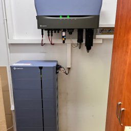 Sun Tech Fotowoltaika Rainczuk Sebastian - Szary magazyn energii Solplanet z podłączonym inwerterem na białej ścianie. Widoczne kable i diody sygnalizacyjne. Fragment szafki po prawej stronie.