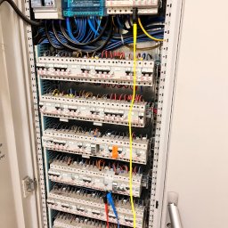 Sun Tech Fotowoltaika Rainczuk Sebastian - Otwarta szafa elektryczna z bezpiecznikami, okablowaniem i miernikiem w trakcie pomiarów. Widoczne przewody pomiarowe w kolorach żółtym, niebieskim i czerwonym.