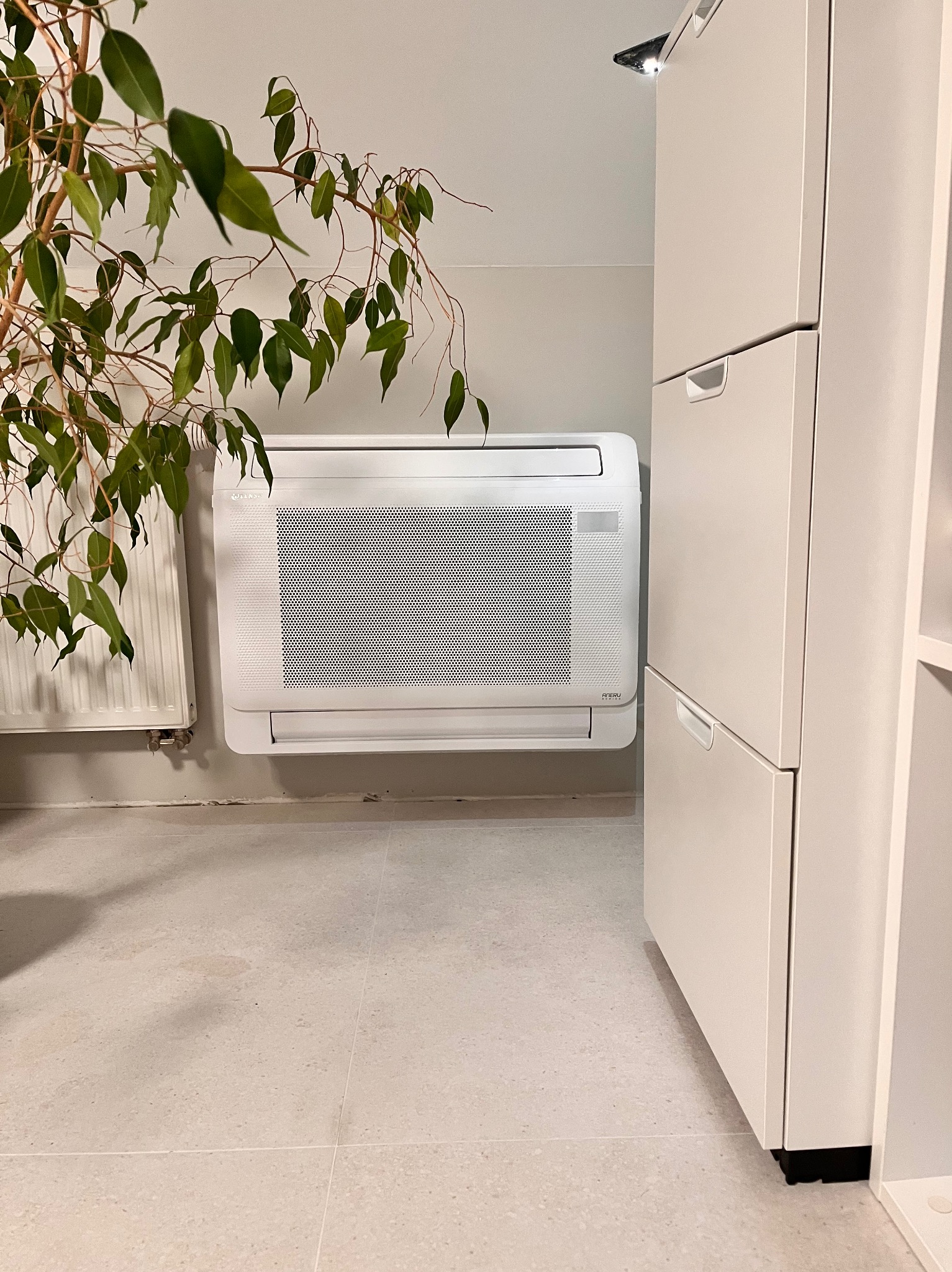 Nowoczesny, biały klimakonwektor pod parapetem, obok szafki na buty i rośliny doniczkowej. Widoczne ślady po instalacji. Neutralne wnętrze.