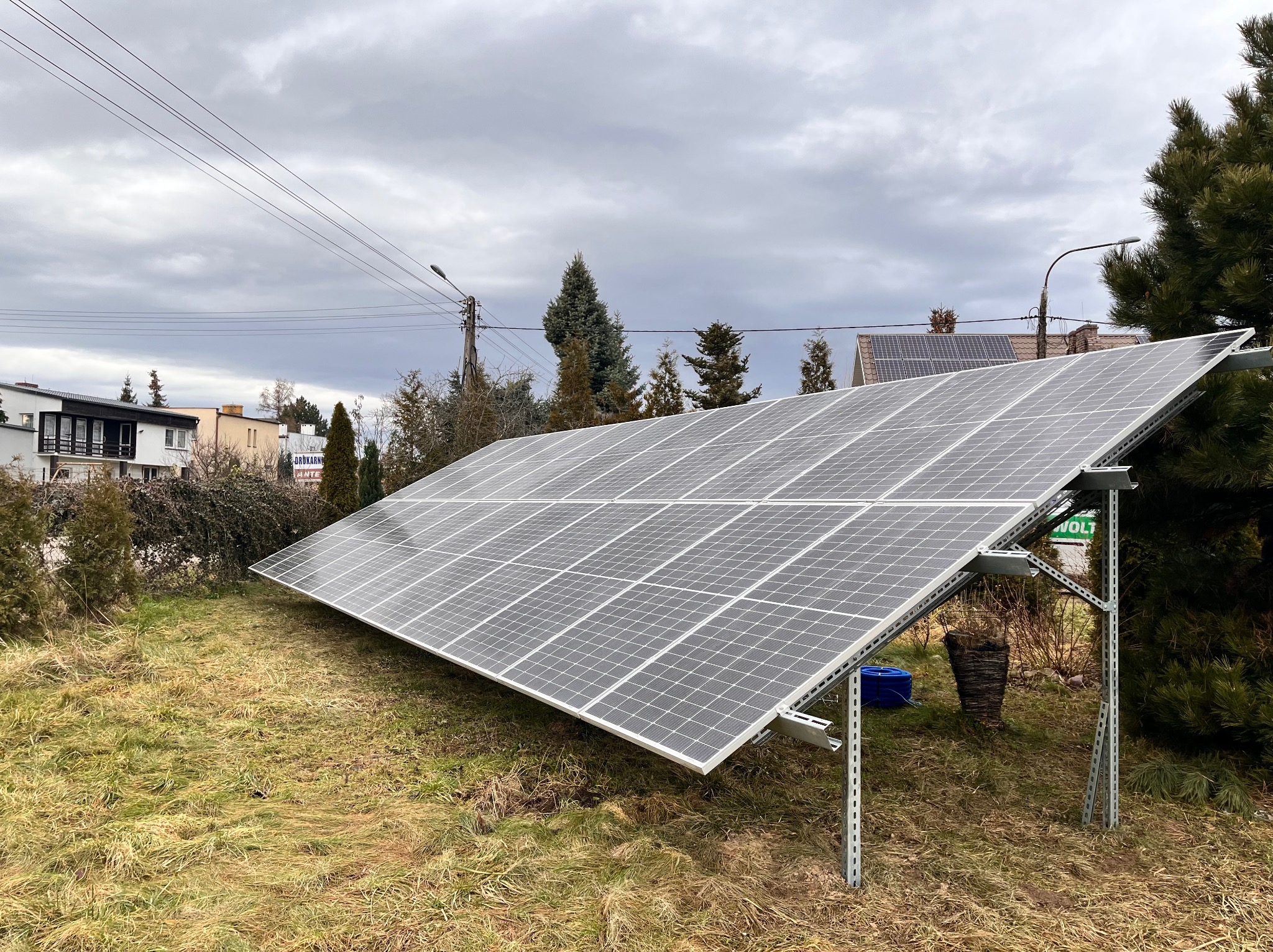 Panel fotowoltaiczny na trawie, zamontowany na metalowej konstrukcji. W tle domy i pochmurne niebo. Widoczny fragment instalacji na dachu sąsiedniego budynku.