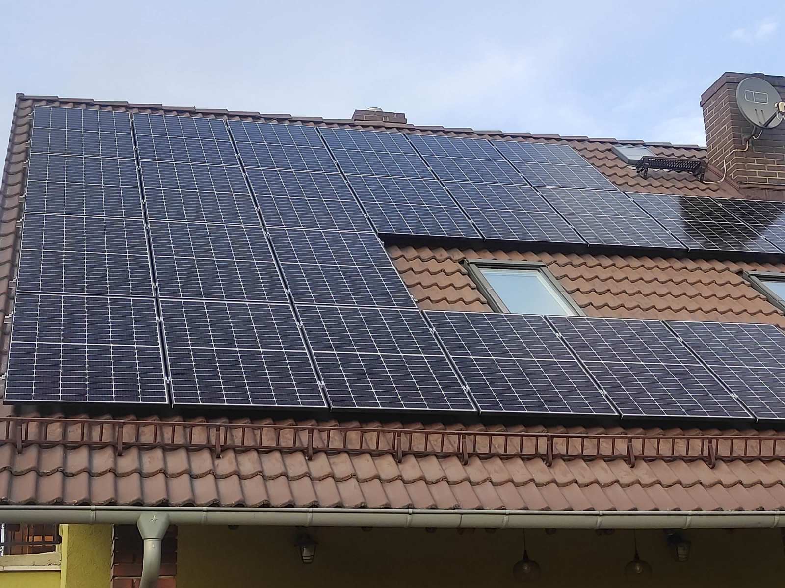 Panele fotowoltaiczne na dachu domu z brązową dachówką, widoczne okna dachowe i komin z anteną satelitarną. Instalacja wkomponowana w istniejącą architekturę.