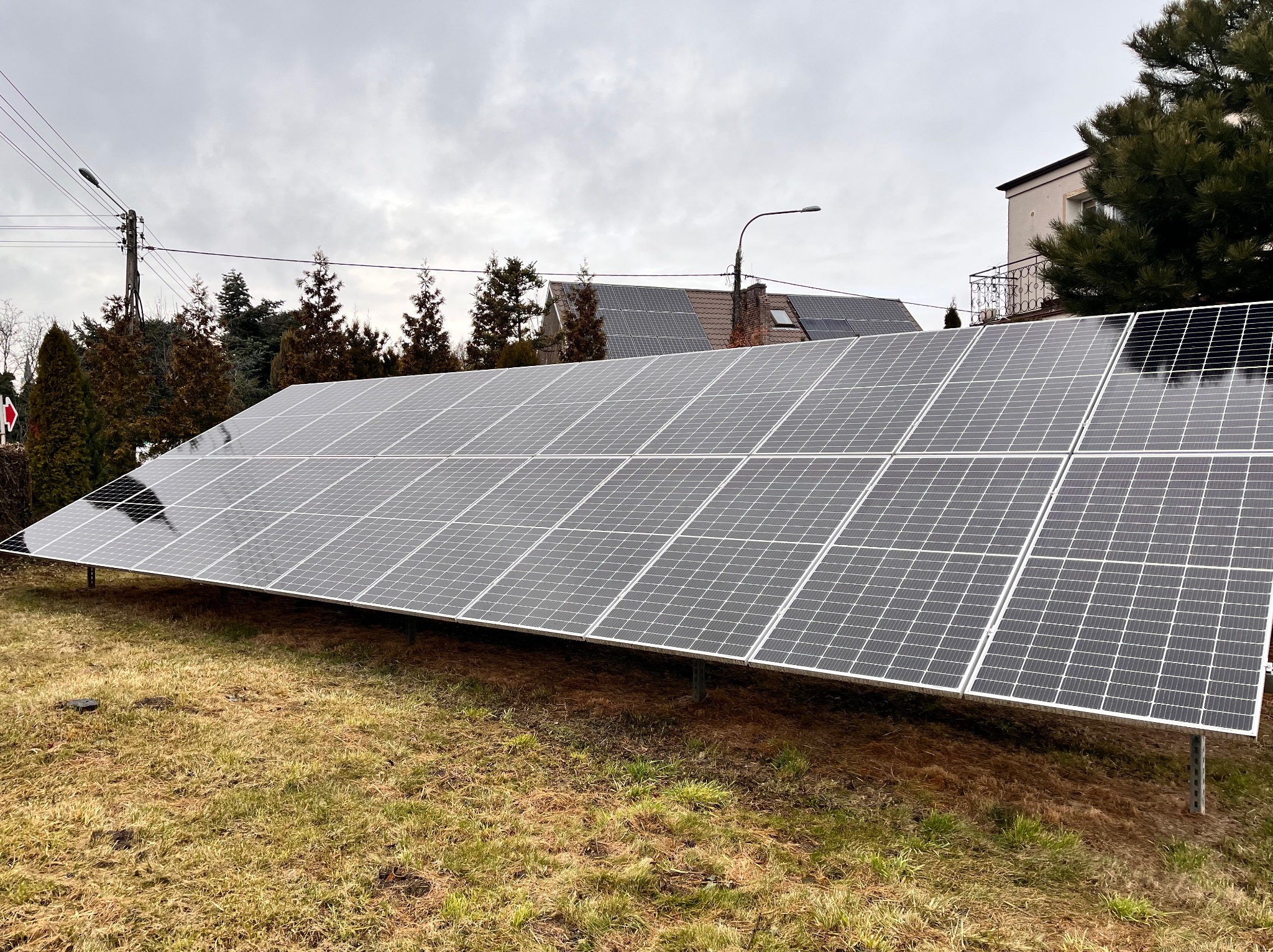 Panel fotowoltaiczny na trawiastym terenie, z domem i drzewami w tle, pochmurne niebo. Widoczne mocowania paneli. Instalacja na gruncie.