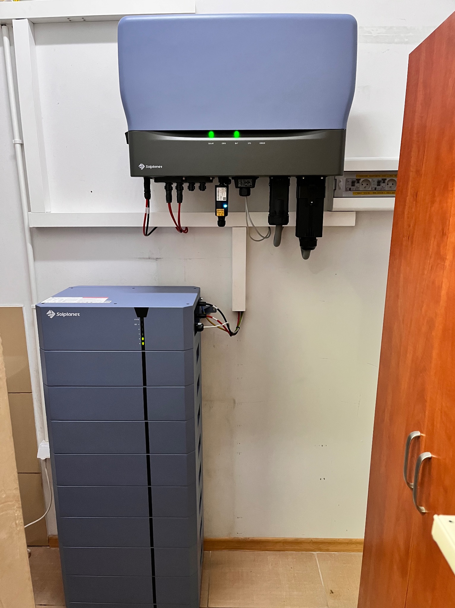 Szary magazyn energii Solplanet z podłączonym inwerterem na białej ścianie. Widoczne kable i diody sygnalizacyjne. Fragment szafki po prawej stronie.