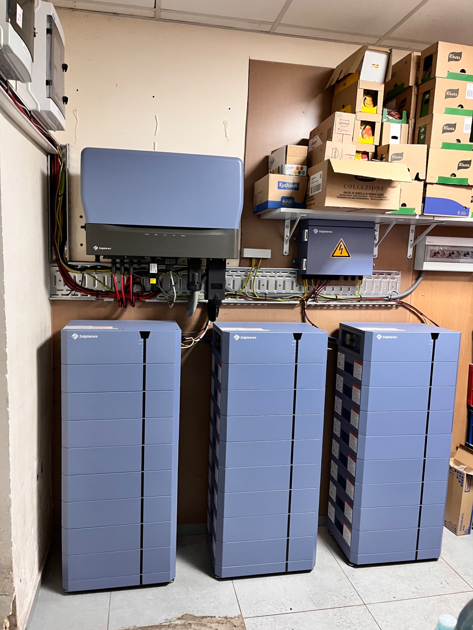Instalacja magazynów energii Solplanet: trzy moduły bateryjne w rzędzie, podłączone do inwertera na ścianie. Widoczne okablowanie i elementy montażowe. Tło stanowią kartony na półce.