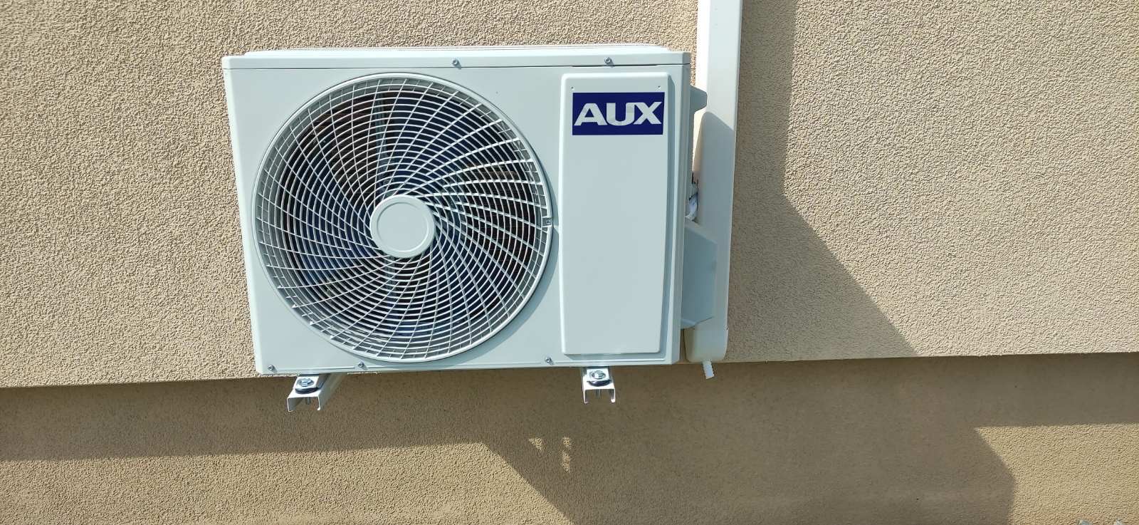Zamontowana na zewnątrz, jasna jednostka klimatyzacji AUX na beżowej, teksturowanej ścianie budynku. Widoczne wsporniki montażowe i fragment rury odprowadzającej.