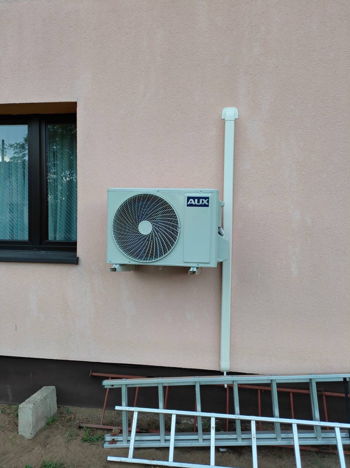 Zamontowana jednostka zewnętrzna klimatyzacji AUX na elewacji budynku, obok okna. Widoczna rura odprowadzająca skropliny i drabina oparta o ścianę.