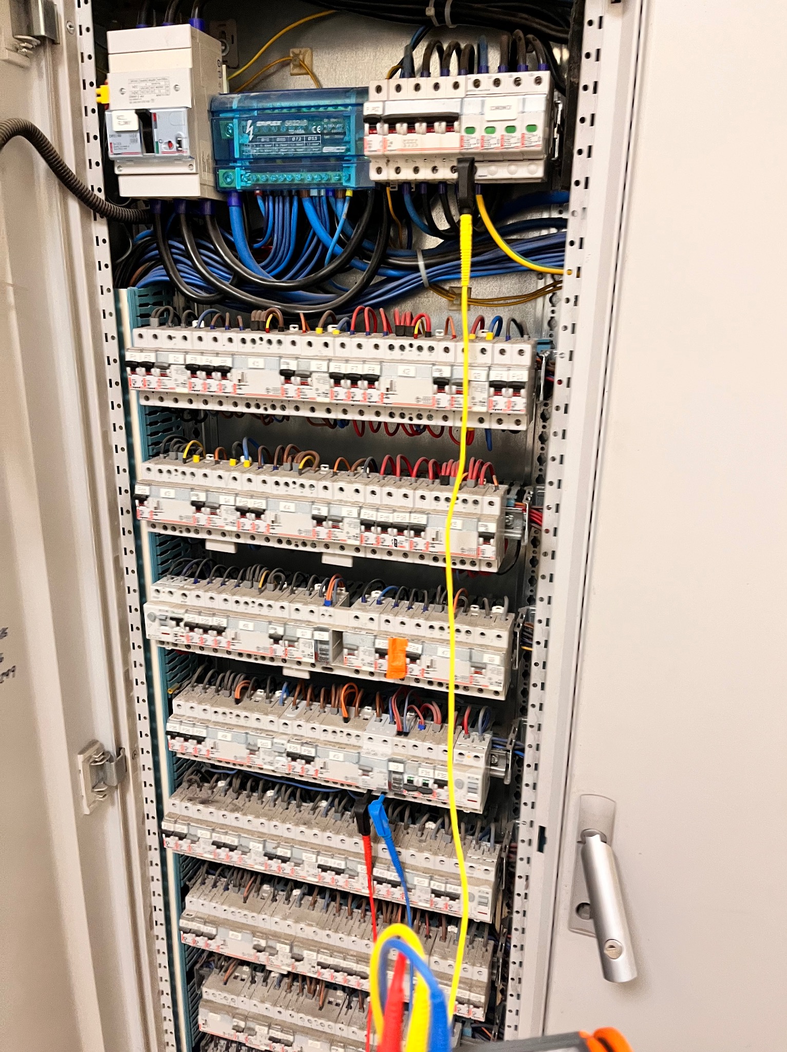 Otwarta szafa elektryczna z bezpiecznikami, okablowaniem i miernikiem w trakcie pomiarów. Widoczne przewody pomiarowe w kolorach żółtym, niebieskim i czerwonym.