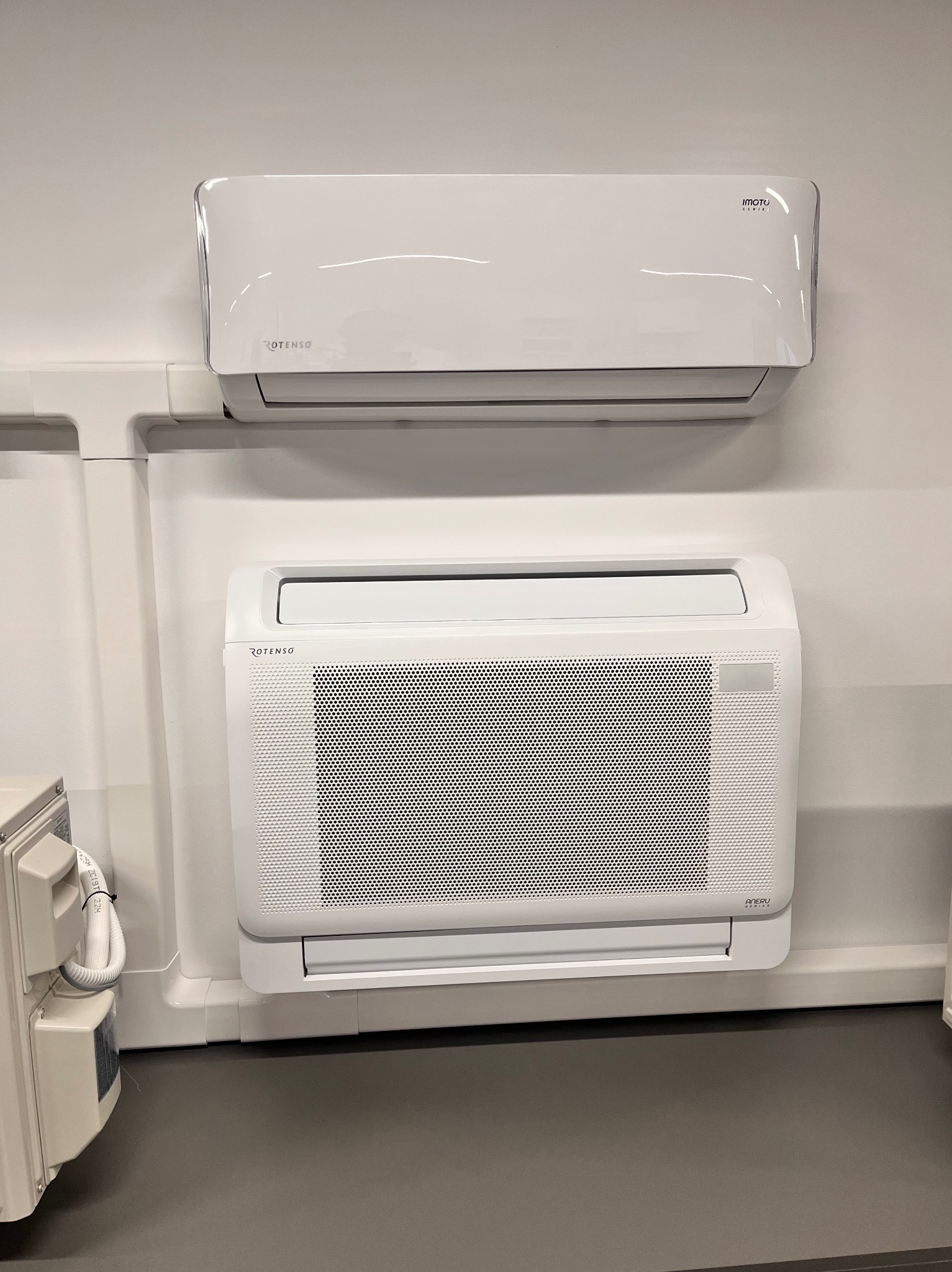 Dwa białe klimatyzatory Rotenso zamontowane na ścianie. Górny model ścienny, dolny przypodłogowy z kratką. Widoczne elementy instalacji.