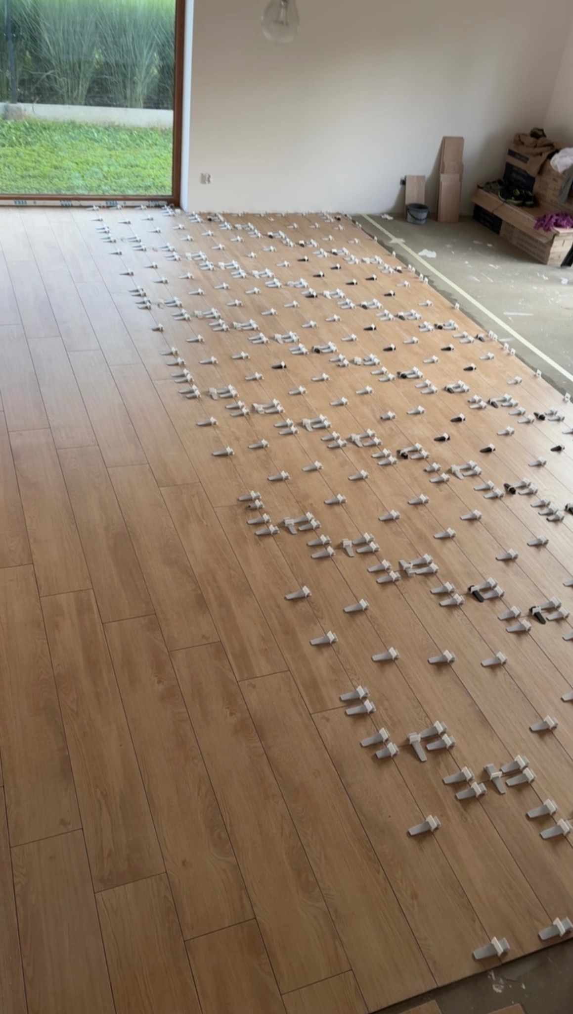 Układanie paneli podłogowych w jasnym wnętrzu. Widoczne białe klipsy dystansowe, zapewniające równomierne odstępy. Częściowo ułożona podłoga z paneli imitujących drewno.