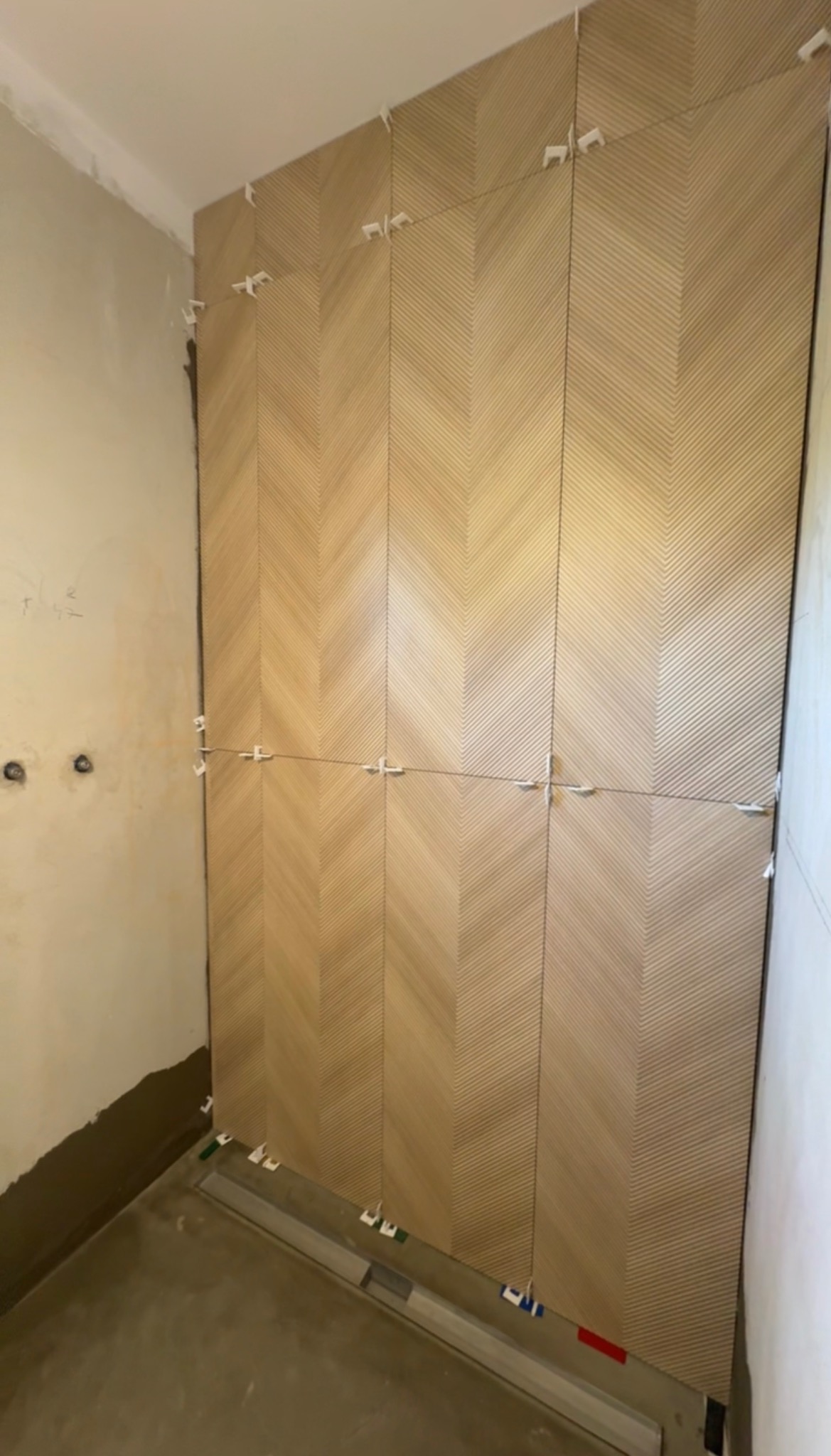 Układanie płytek w jodełkę na ścianie w łazience. Widoczne białe krzyżyki dystansowe i poziomica. Częściowo wykończona ściana, surowy stan podłogi i ściany obok.