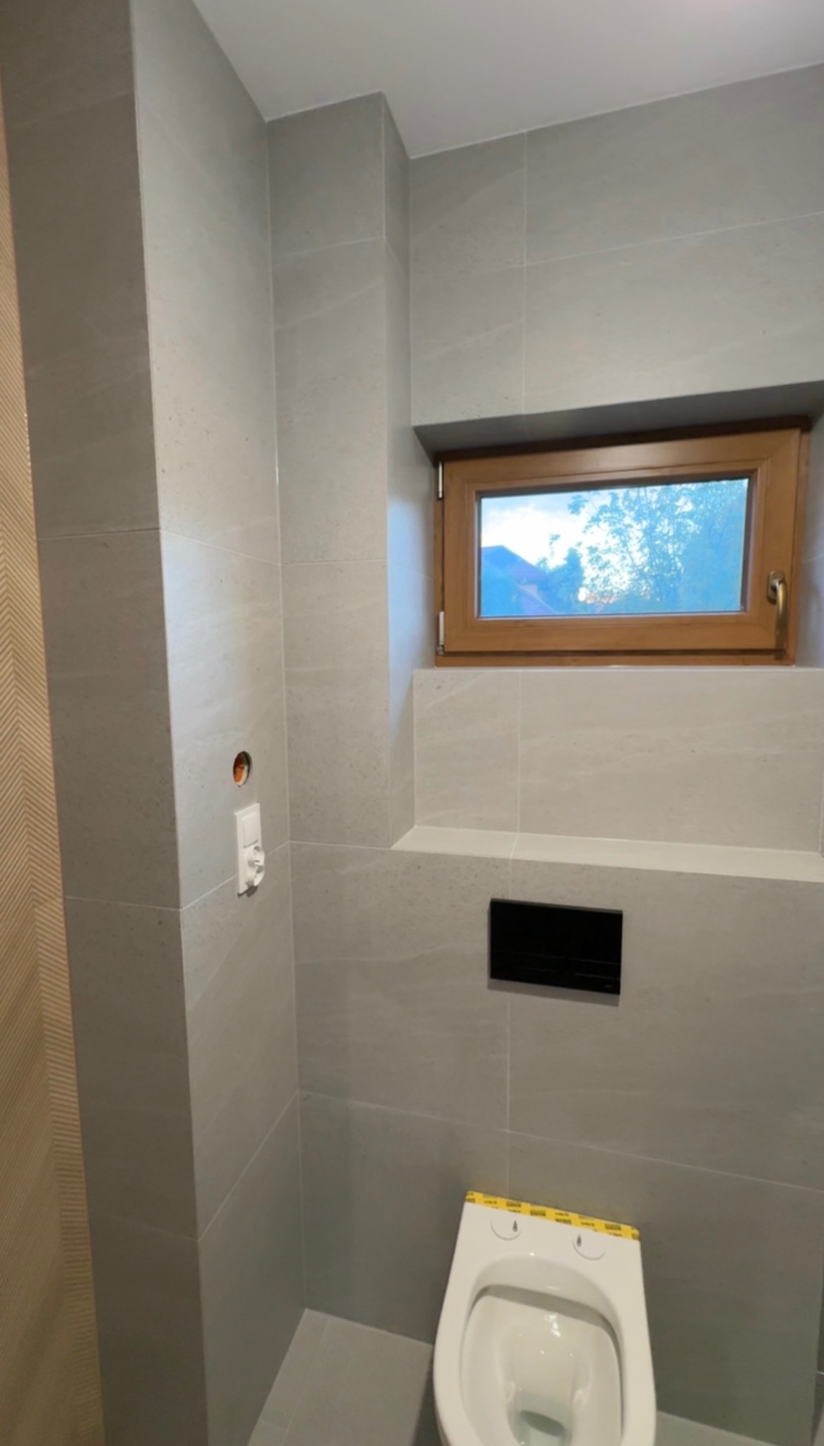 Wykończona łazienka z szarymi płytkami, małym oknem w drewnianej ramie i podwieszaną toaletą z żółtą taśmą zabezpieczającą. Widoczne gniazdko i czarny przycisk spłuczki.