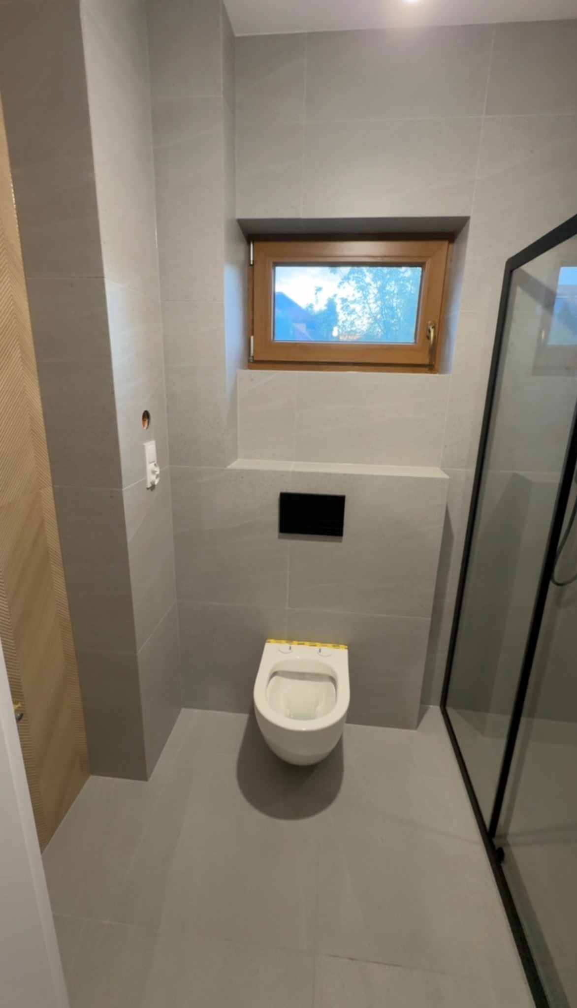 Nowoczesna łazienka w szarościach z podwieszaną toaletą, drewnianym oknem i kabiną prysznicową z czarnymi ramami. Minimalistyczny design, płytki imitujące kamień.