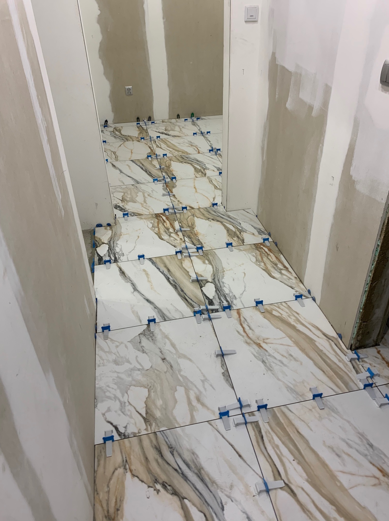 Układanie płytek imitujących marmur na podłodze w korytarzu. Widoczne niebieskie klipsy poziomujące. Ściany przygotowane do malowania.