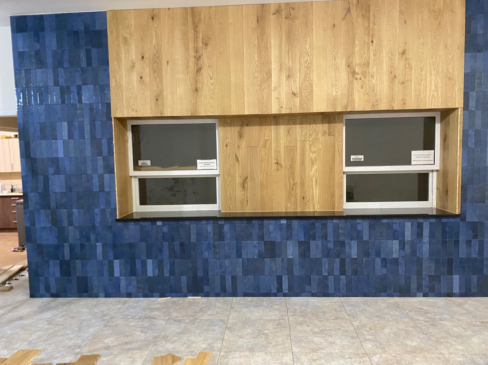 Niebieska ściana z płytek w pionowe pasy, drewniana zabudowa z dwoma oknami, jasna podłoga z płytek. Widoczne fragmenty paneli podłogowych.
