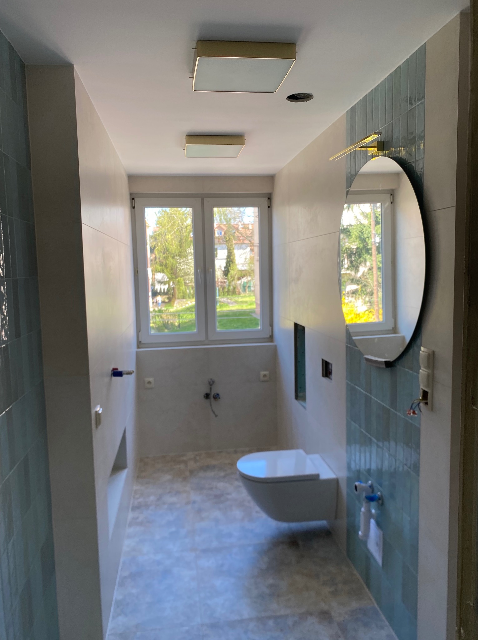 Nowoczesna łazienka po remoncie: błękitna glazura, podwieszana toaleta, okrągłe lustro i okno z widokiem na zieleń. Minimalistyczne oświetlenie sufitowe.