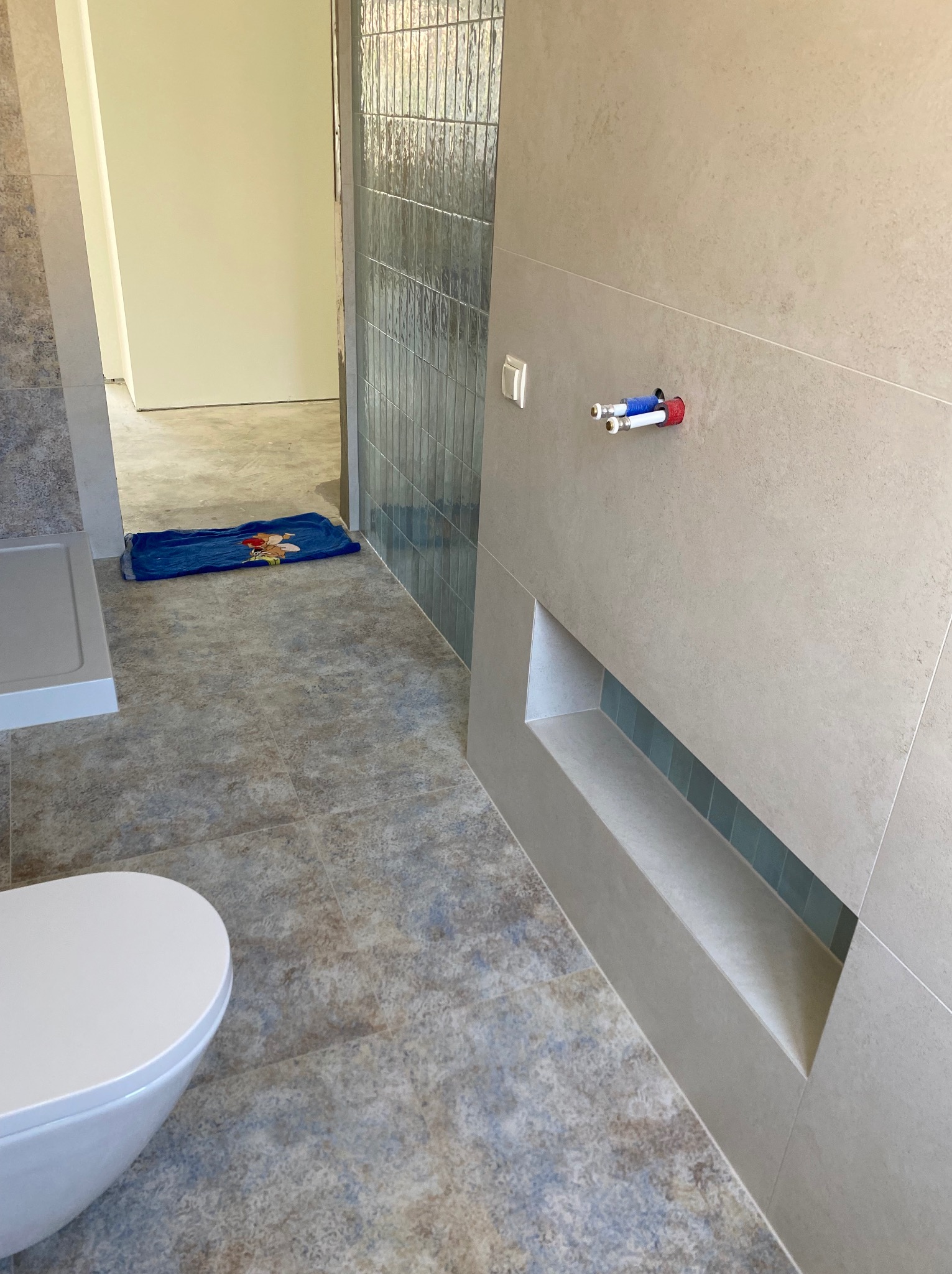 Wykończona łazienka z beżowymi płytkami, wnęką z niebieskimi akcentami i widocznymi rurami. Na podłodze płytki imitujące kamień i fragment toalety.