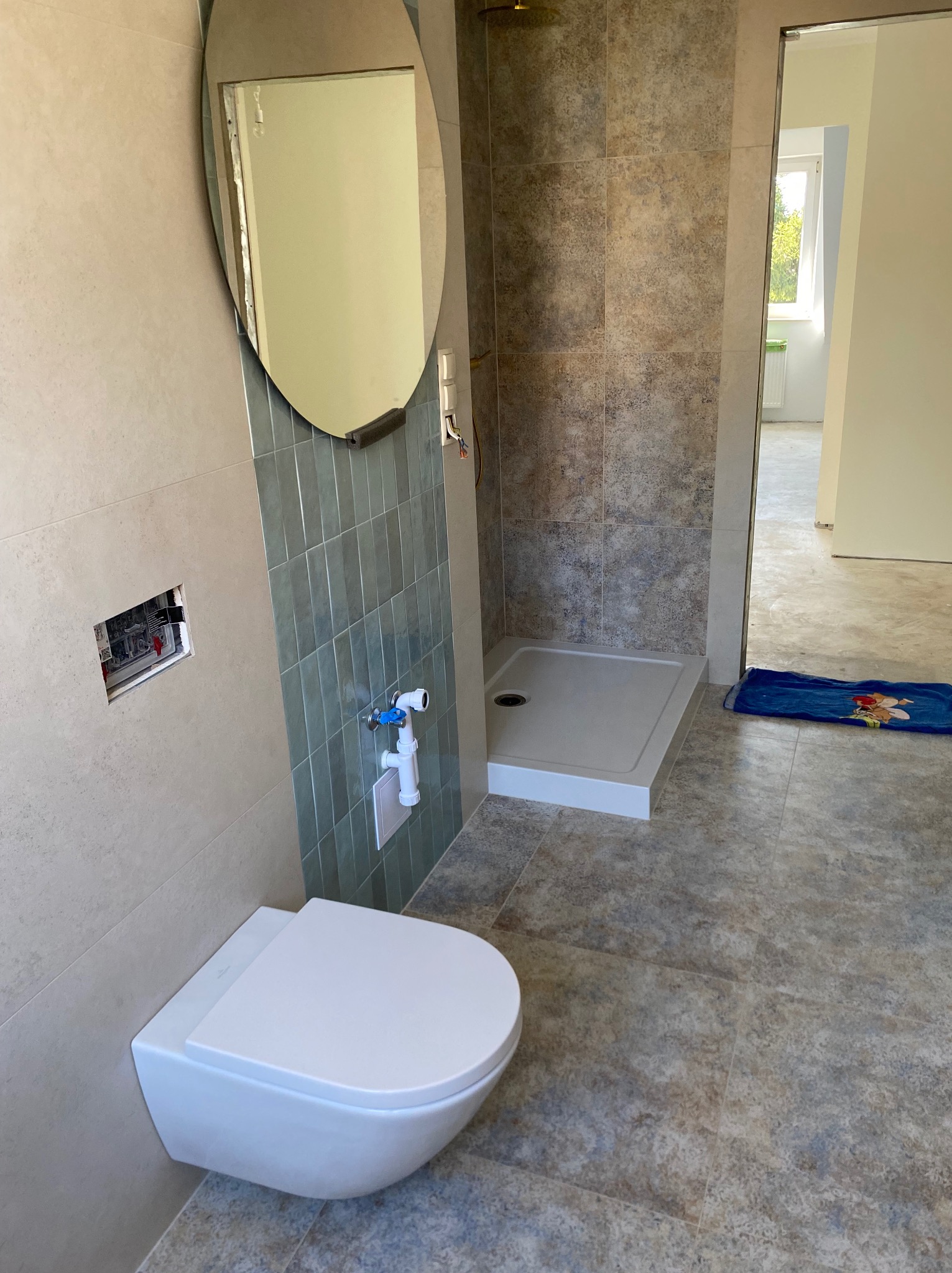 Nowoczesna łazienka po remoncie: podwieszana toaleta, kabina prysznicowa, owalne lustro. Widoczne płytki w odcieniach beżu i szarości. Otwór w ścianie przed zabudową.