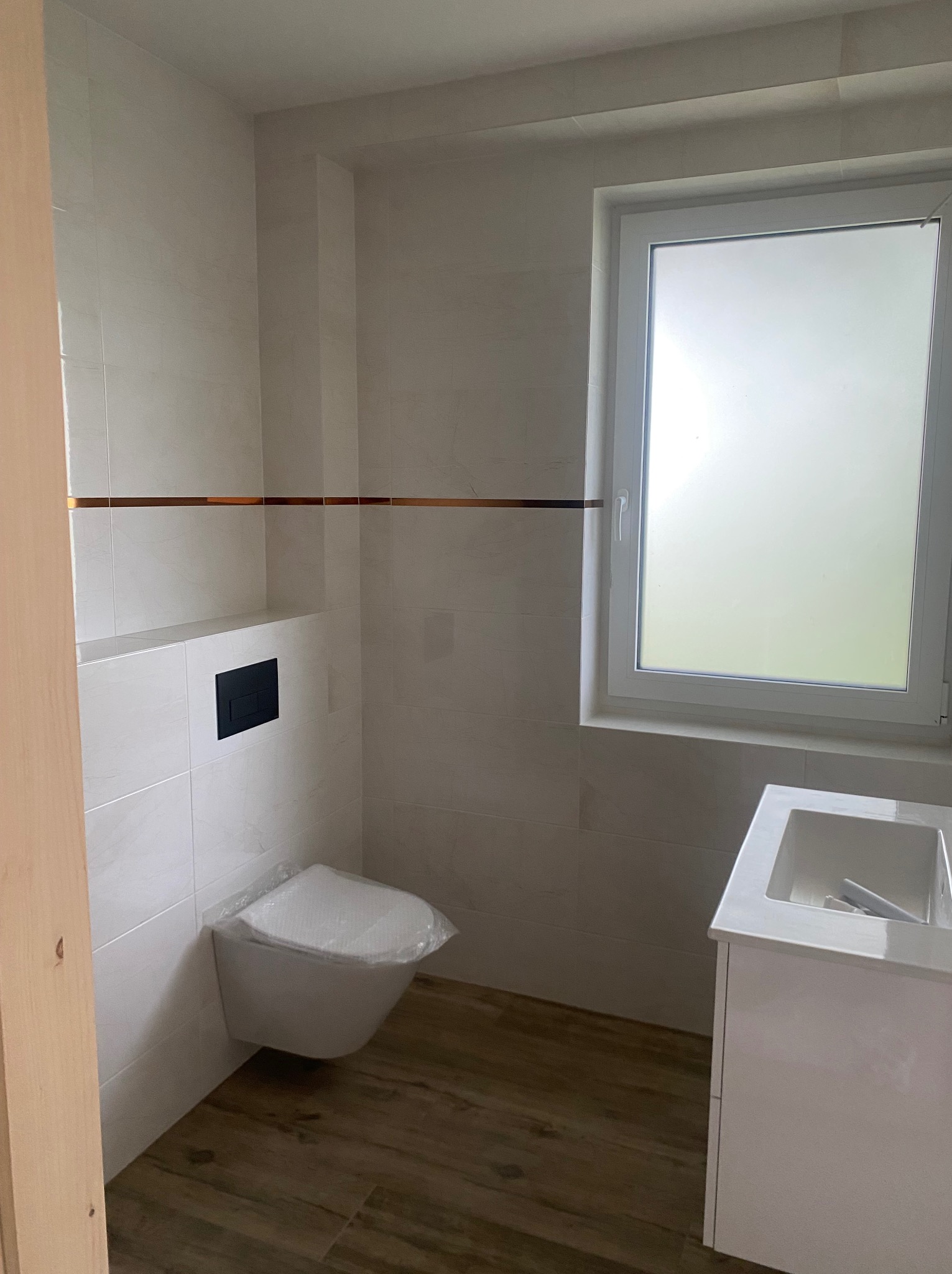 Nowoczesna łazienka po remoncie: białe płytki z miedzianym akcentem, podwieszana toaleta z czarnym panelem, drewnopodobne panele podłogowe i biała szafka z umywalką.