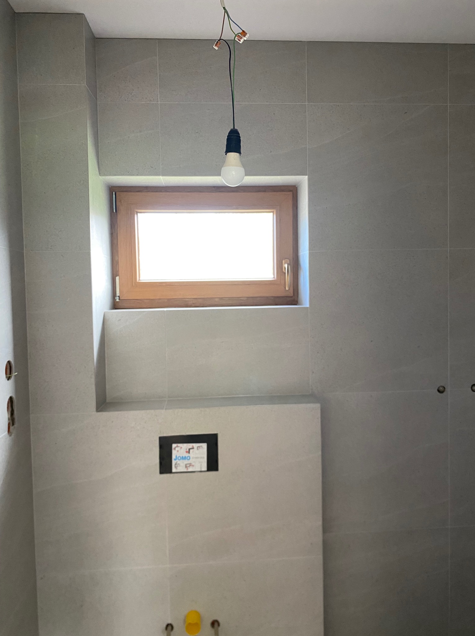 Jasne, matowe płytki w łazience z oknem w drewnianej ramie. Widoczna instalacja podtynkowa WC i prowizoryczne oświetlenie żarówką.