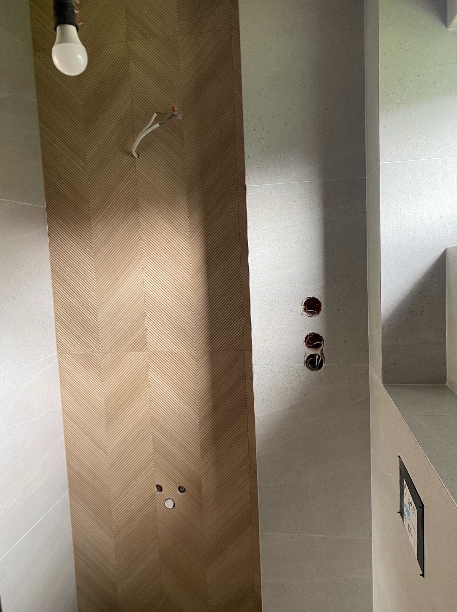 Wykończona ściana w łazience z dekoracyjną płytką w jodełkę i przygotowanymi punktami elektrycznymi. Widoczne szare płytki i surowa żarówka.