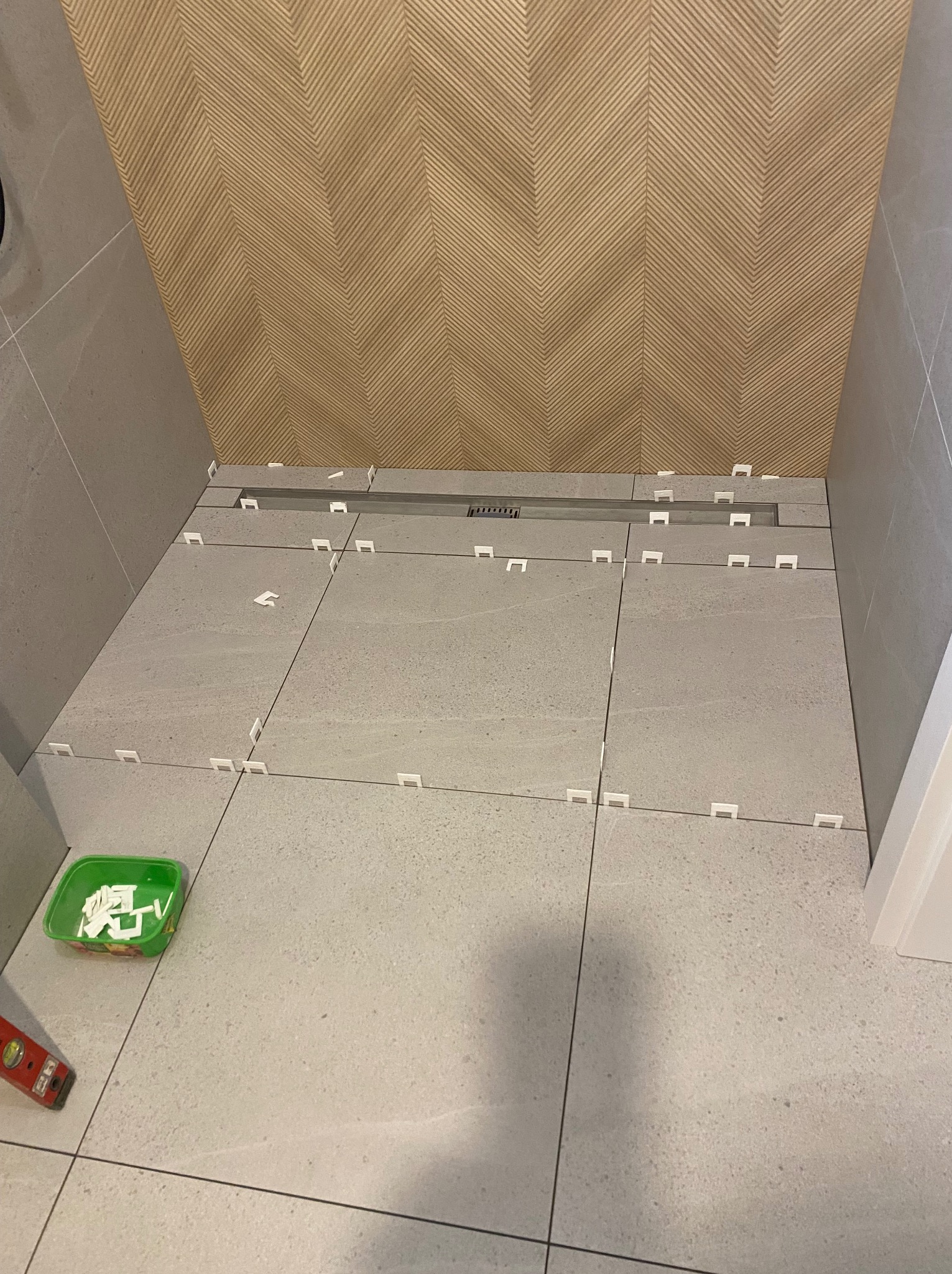 Układanie szarych płytek podłogowych z użyciem białych krzyżyków dystansowych. W tle ściana z dekoracyjną okładziną w jodełkę. Widoczny odpływ liniowy.