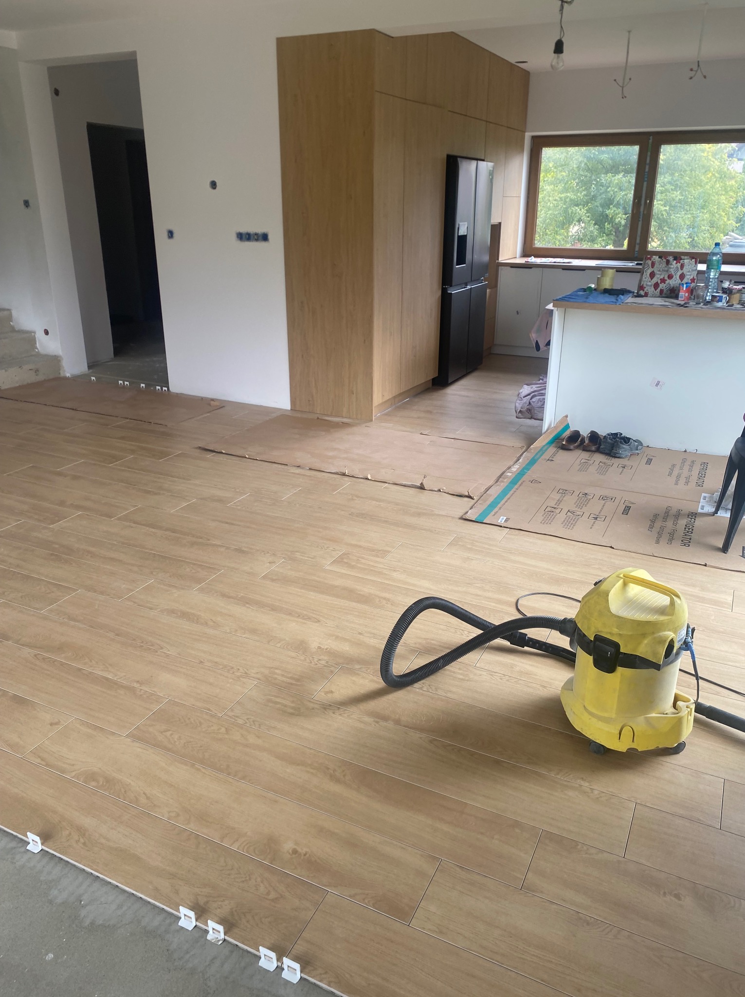 Układanie płytek podłogowych w jasnym odcieniu drewna, z widocznym odkurzaczem przemysłowym i elementami montażowymi. Częściowo wykończone pomieszczenie z zabudową kuchenną.