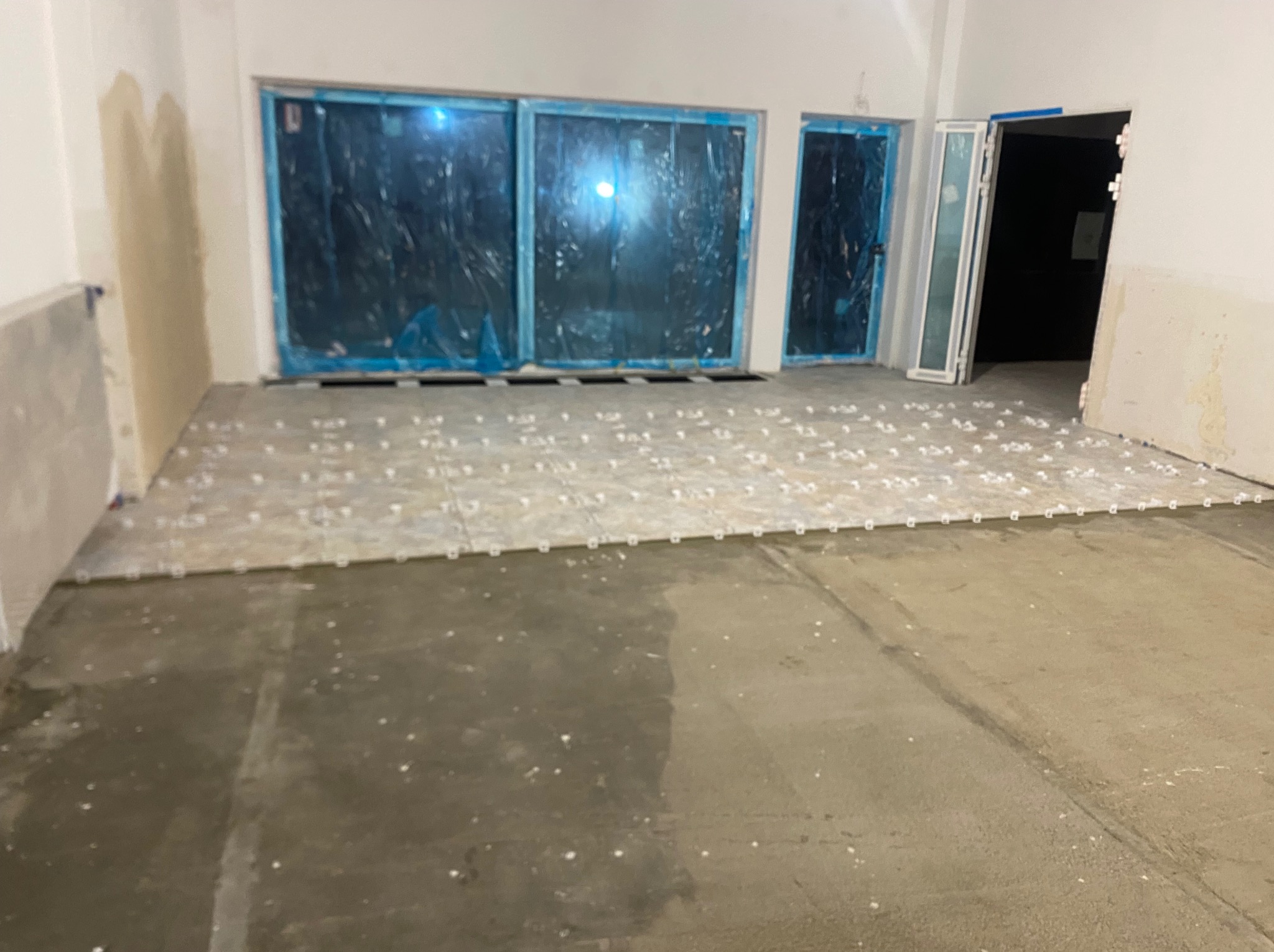 Przygotowanie do układania płytek podłogowych w pomieszczeniu z zabezpieczonymi folią oknami i drzwiami. Widoczne krzyżyki dystansowe.