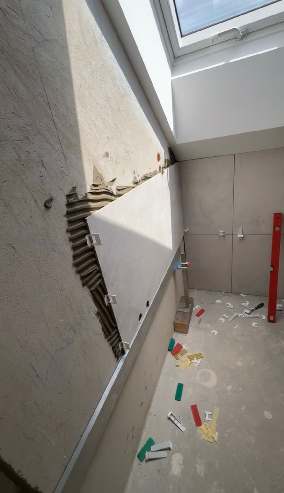 Układanie jasnych płytek na ścianie w łazience, widoczne narzędzia i poziomica. Częściowo wykończona ściana, płytki z dystansami. Okno dachowe w tle.