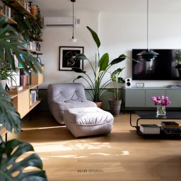 MAR Studio Małgorzata Rembowska - Jasne wnętrze salonu z drewnianą podłogą, szarą sofą modułową, roślinami doniczkowymi i regałem z książkami. Minimalistyczny design z akcentami zieleni.