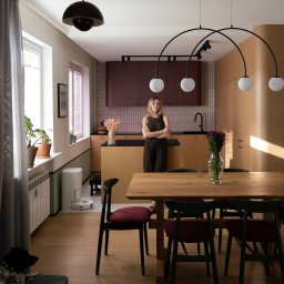 MAR Studio Małgorzata Rembowska - Jasne wnętrze z drewnianym stołem i krzesłami. W tle kobieta na tle kuchni z bordowymi szafkami. Uwagę przykuwa lampa łukowa nad stołem i robot sprzątający w rogu.