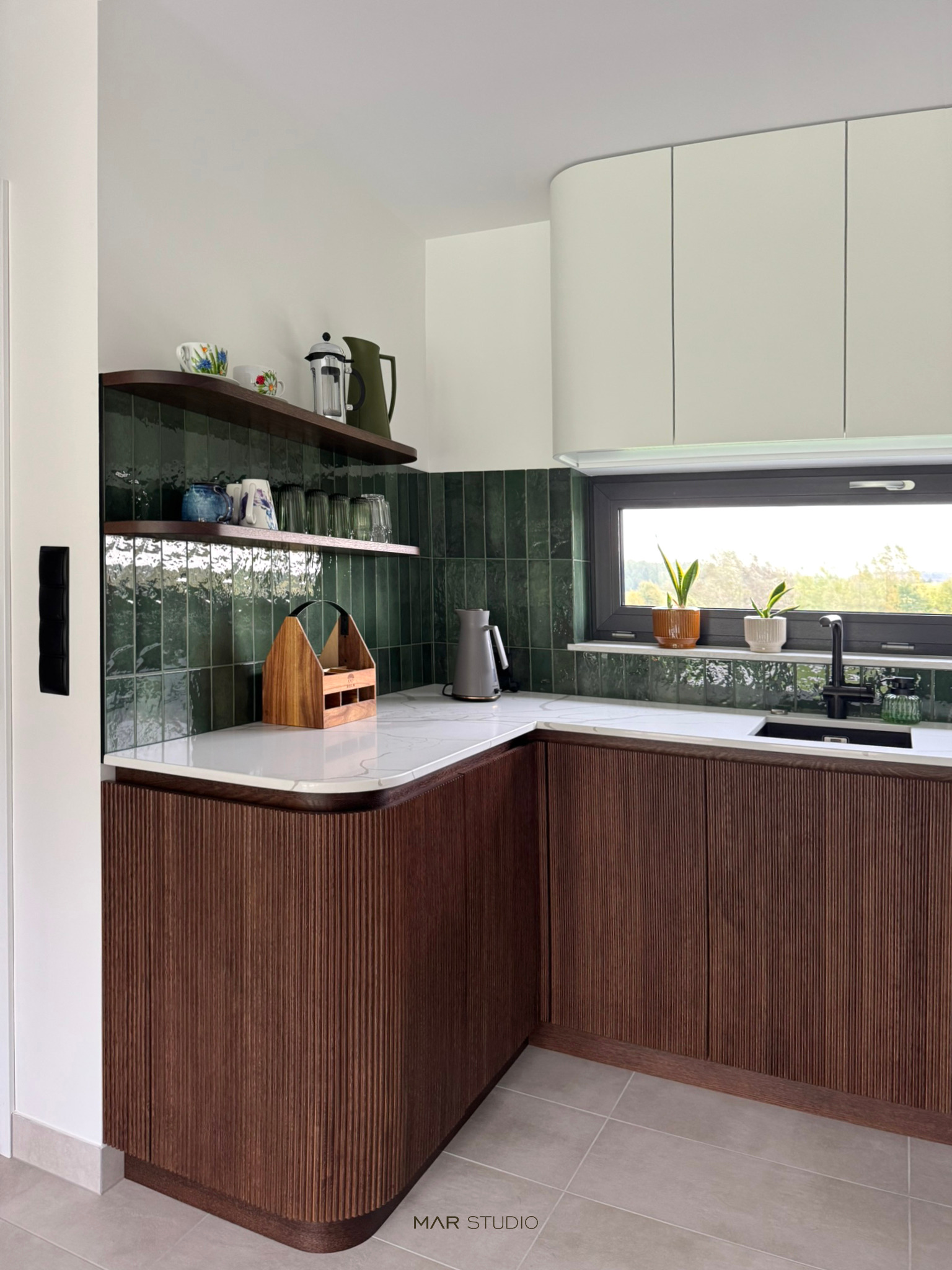 Nowoczesna kuchnia z ciemnozielonymi płytkami i drewnianymi szafkami z ryflowanym frontem. Na blacie marmur, a na półkach i parapecie dekoracje.