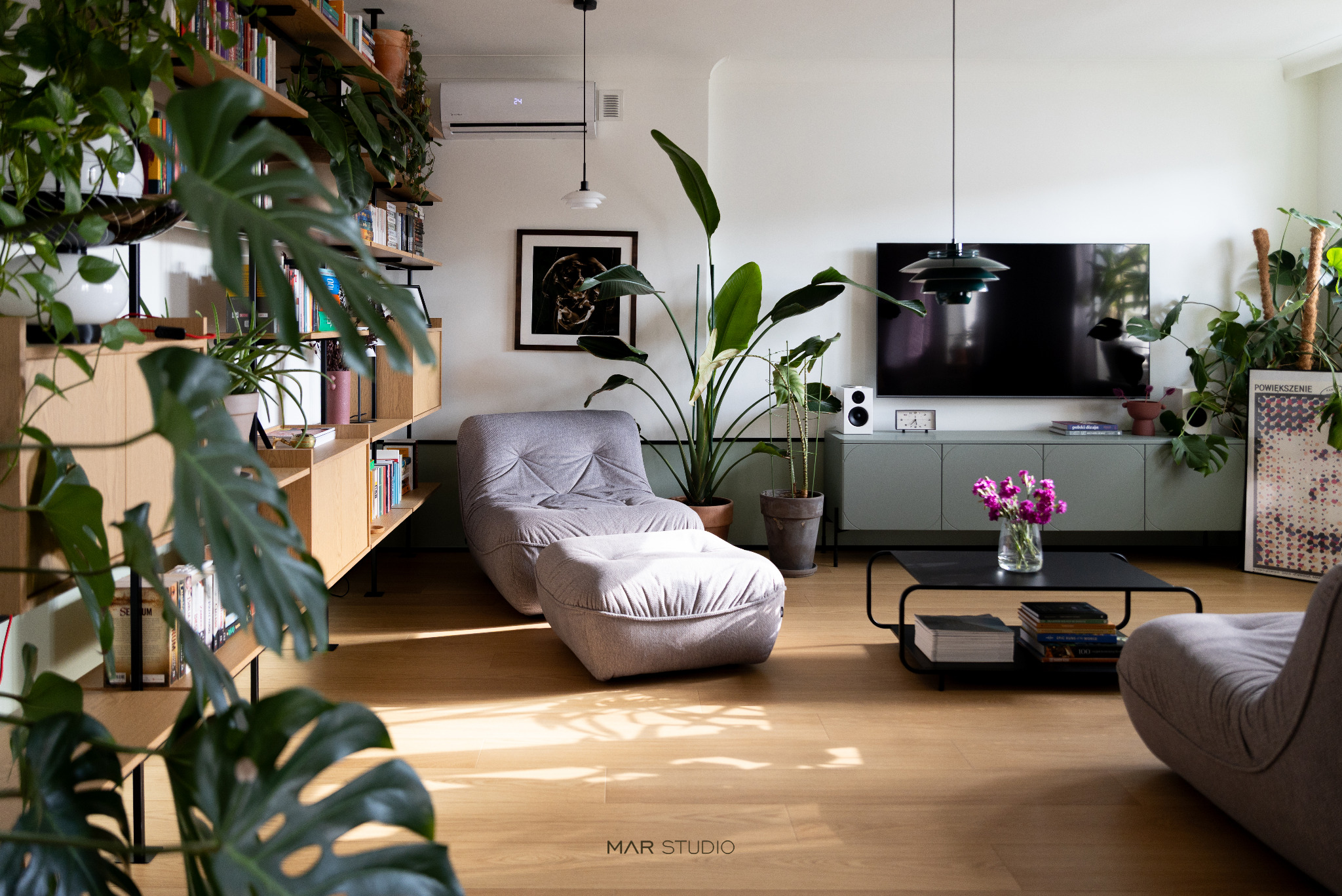 Jasne wnętrze salonu z drewnianą podłogą, szarą sofą modułową, roślinami doniczkowymi i regałem z książkami. Minimalistyczny design z akcentami zieleni.