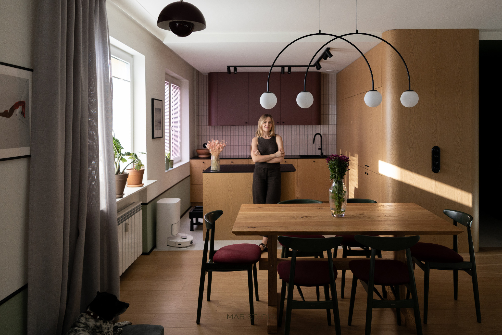 Jasne wnętrze z drewnianym stołem i krzesłami. W tle kobieta na tle kuchni z bordowymi szafkami. Uwagę przykuwa lampa łukowa nad stołem i robot sprzątający w rogu.