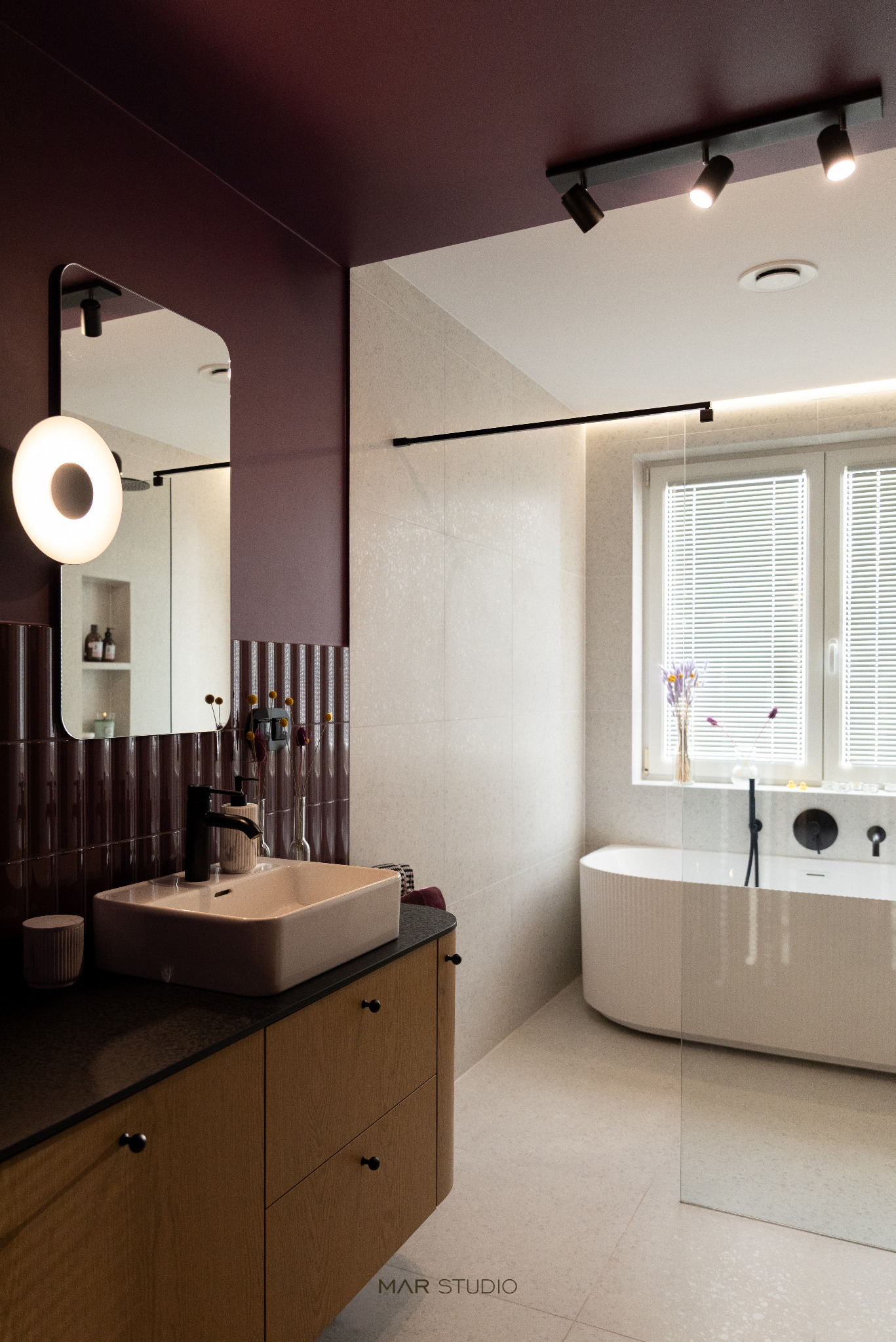 Nowoczesna łazienka z burgundowymi akcentami: umywalka nablatowa, lustro z oświetleniem, wanna za szklaną ścianką. Minimalistyczny design, eleganckie wykończenie.