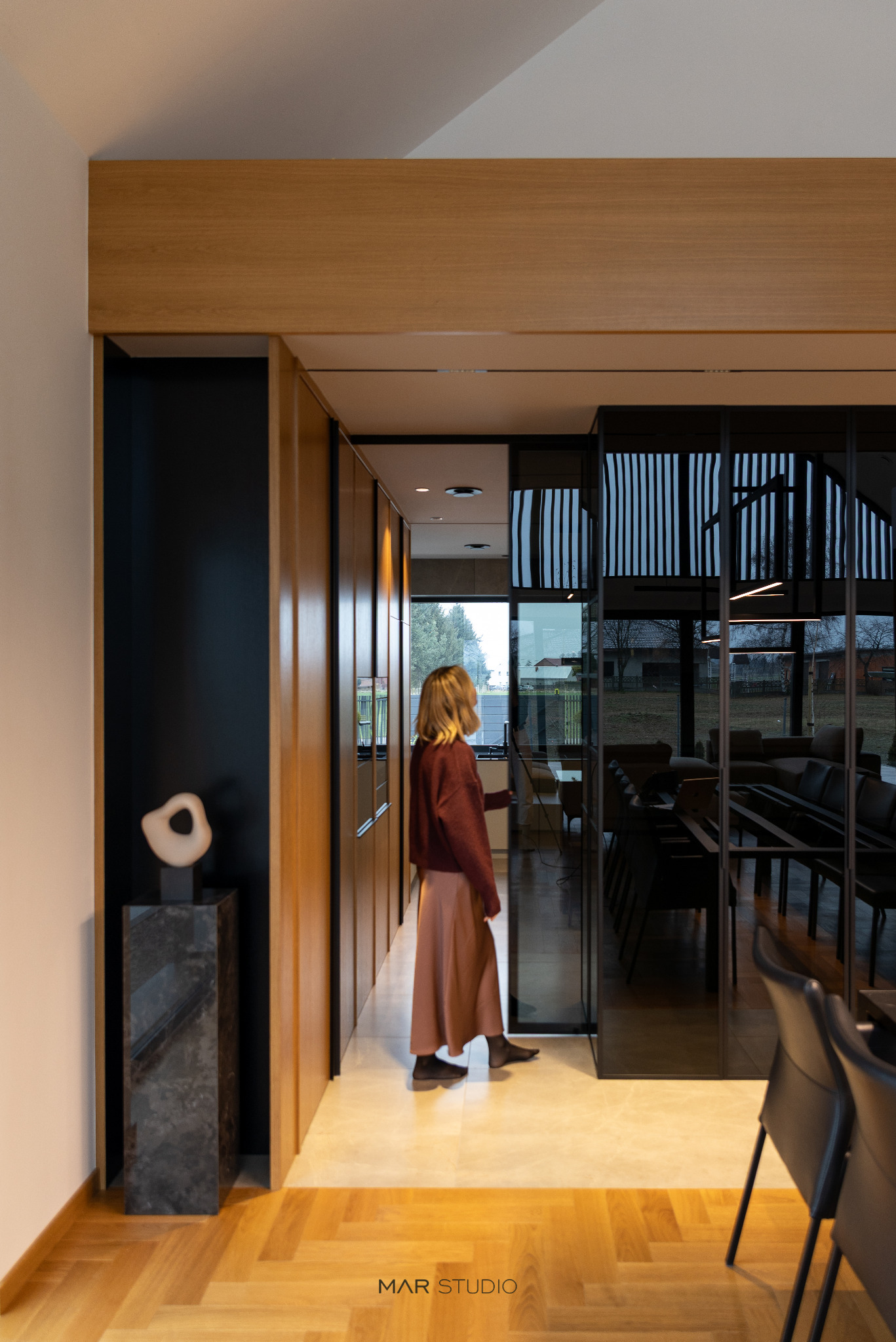 Nowoczesne wnętrze domu z drewnianą podłogą w jodełkę, minimalistyczną rzeźbą na postumencie i przeszkloną ścianą oddzielającą jadalnię. Osoba w tle.