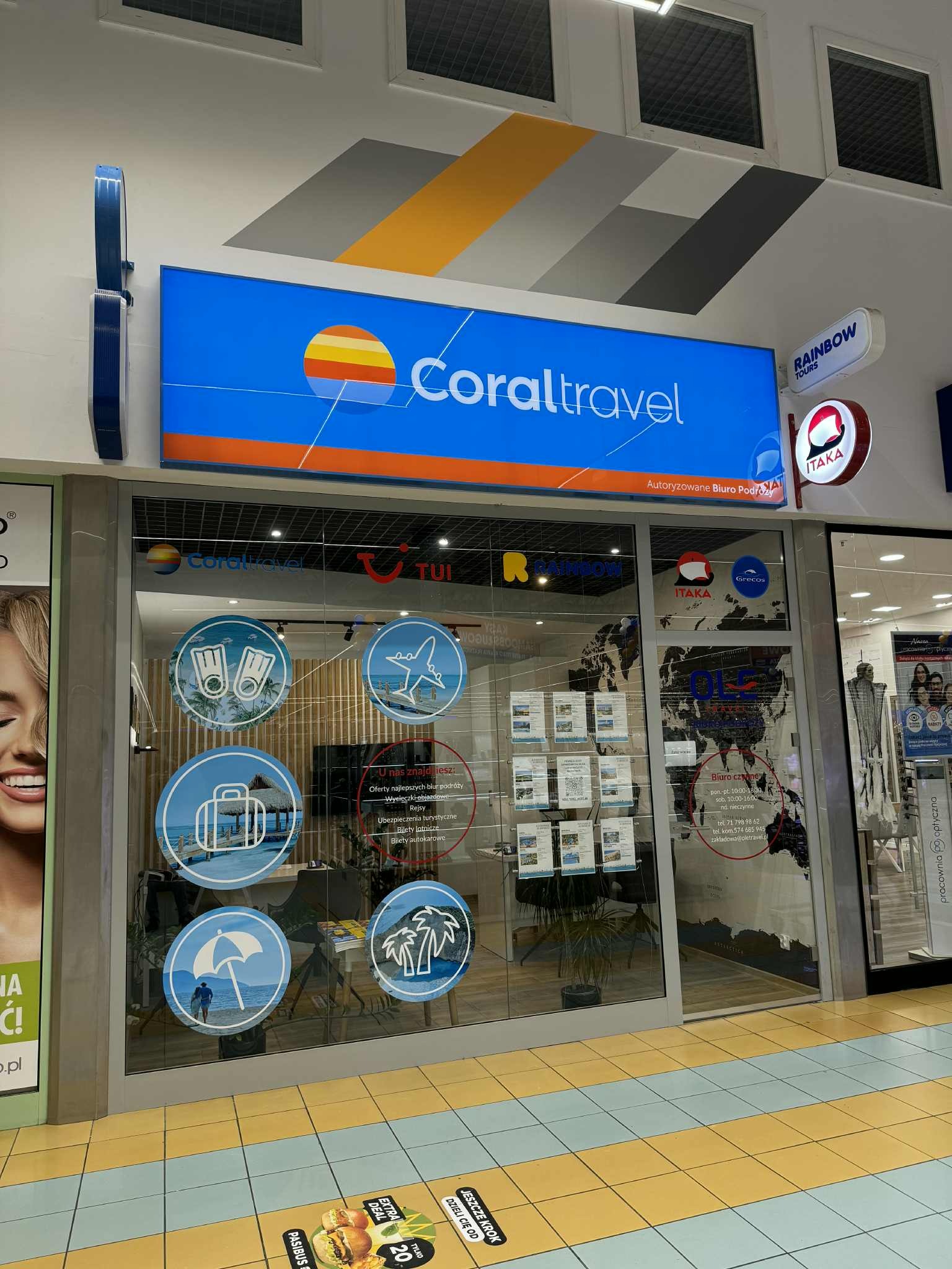 Fasada biura podróży Coral Travel w centrum handlowym, z logo i naklejkami tematycznymi na szybie. Widoczne logo TUI, Rainbow Tours i Itaka. Jasne oświetlenie.