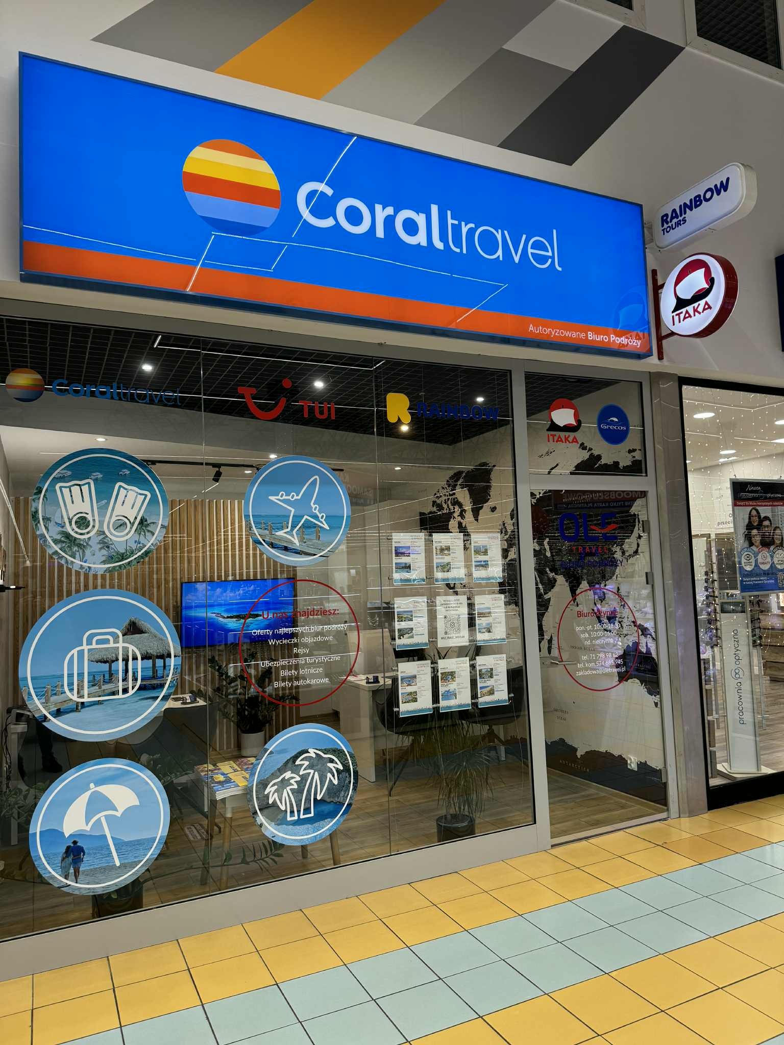 Wejście do biura podróży Coral Travel z logo i naklejkami tematycznymi na szybie, obok loga Rainbow Tours i Itaka. Widoczna mapa świata i oferty wycieczek.