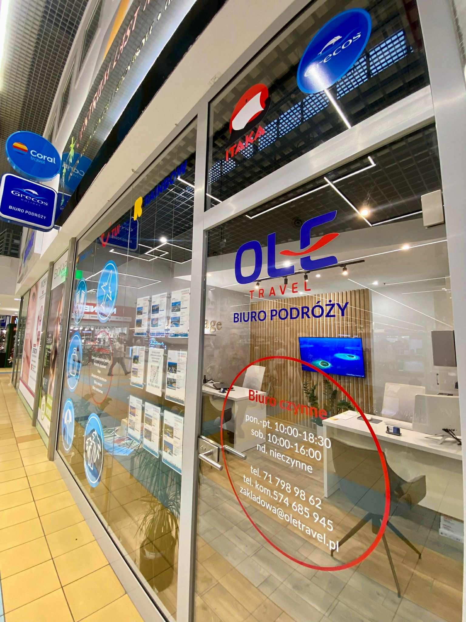 Wejście do biura podróży OLE Travel z logo i informacjami o godzinach otwarcia. Widoczne plakaty z ofertami wycieczek i logotypy Coral Travel i Grecos Holiday.