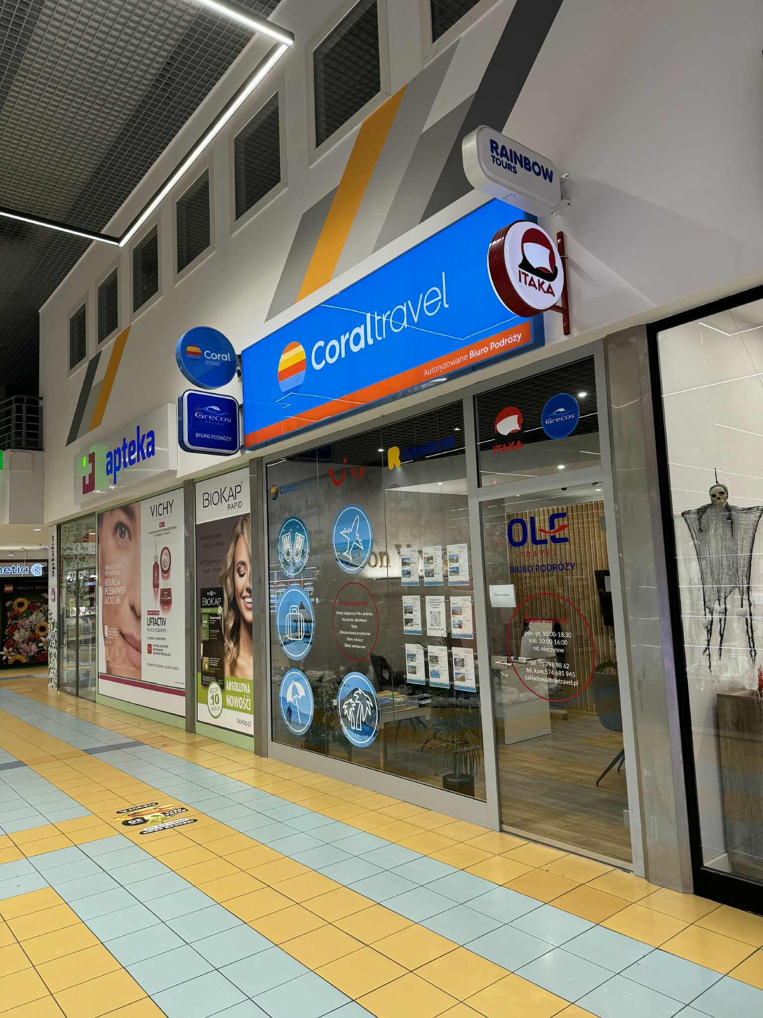 Fasada biura podróży Coral Travel w galerii handlowej, z logo Itaka i Rainbow Tours. Widoczne reklamy apteki i kosmetyków obok.