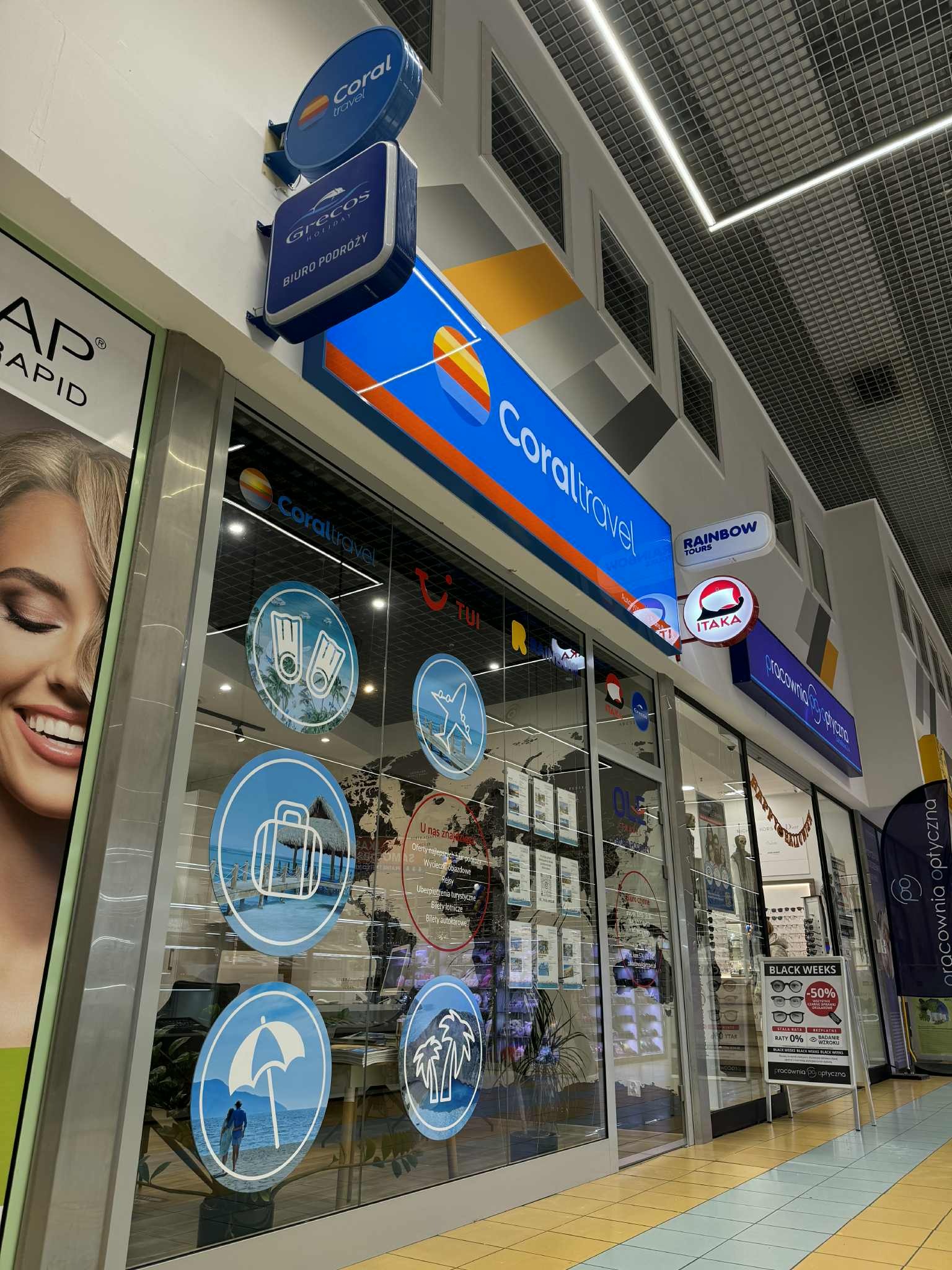Fasada biura podróży Coral Travel w galerii handlowej, z logo i ofertami wycieczek na szybach. Widoczne loga innych biur: Grecos, Itaka, Rainbow Tours.