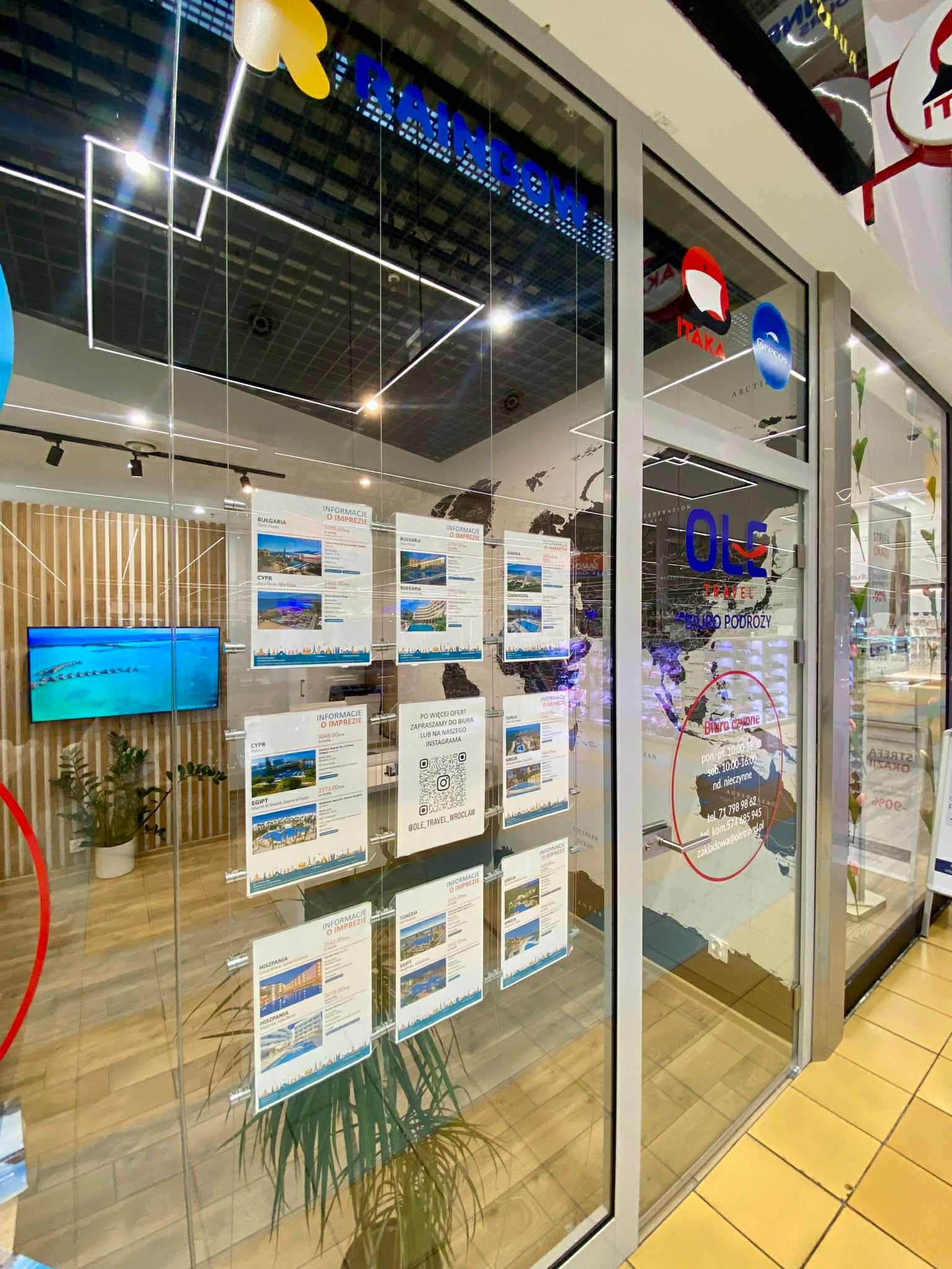 Przeszklona witryna biura podróży z plakatami ofert wycieczek do Bułgarii, Cypru, Egiptu i Hiszpanii. Widoczne logo biura i mapa świata w tle.