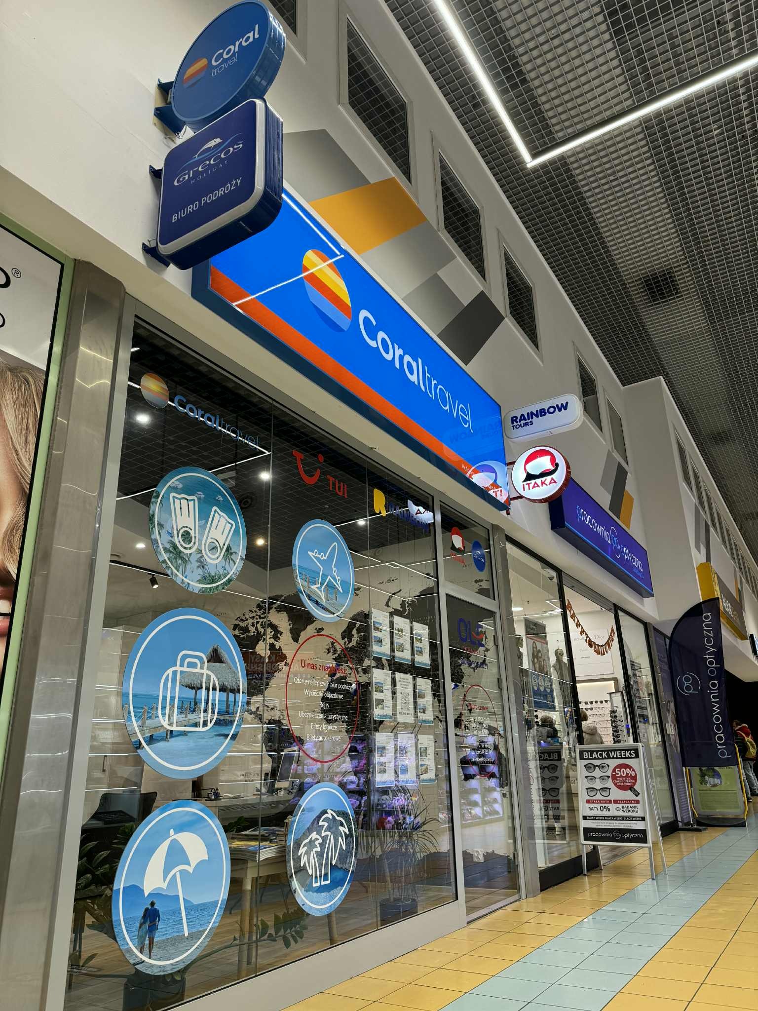 Fasada biura podróży Coral Travel w galerii handlowej, z logo i naklejkami tematycznymi na szybach. Widoczne też loga innych biur: Grecos, Rainbow Tours, Itaka i TUI.