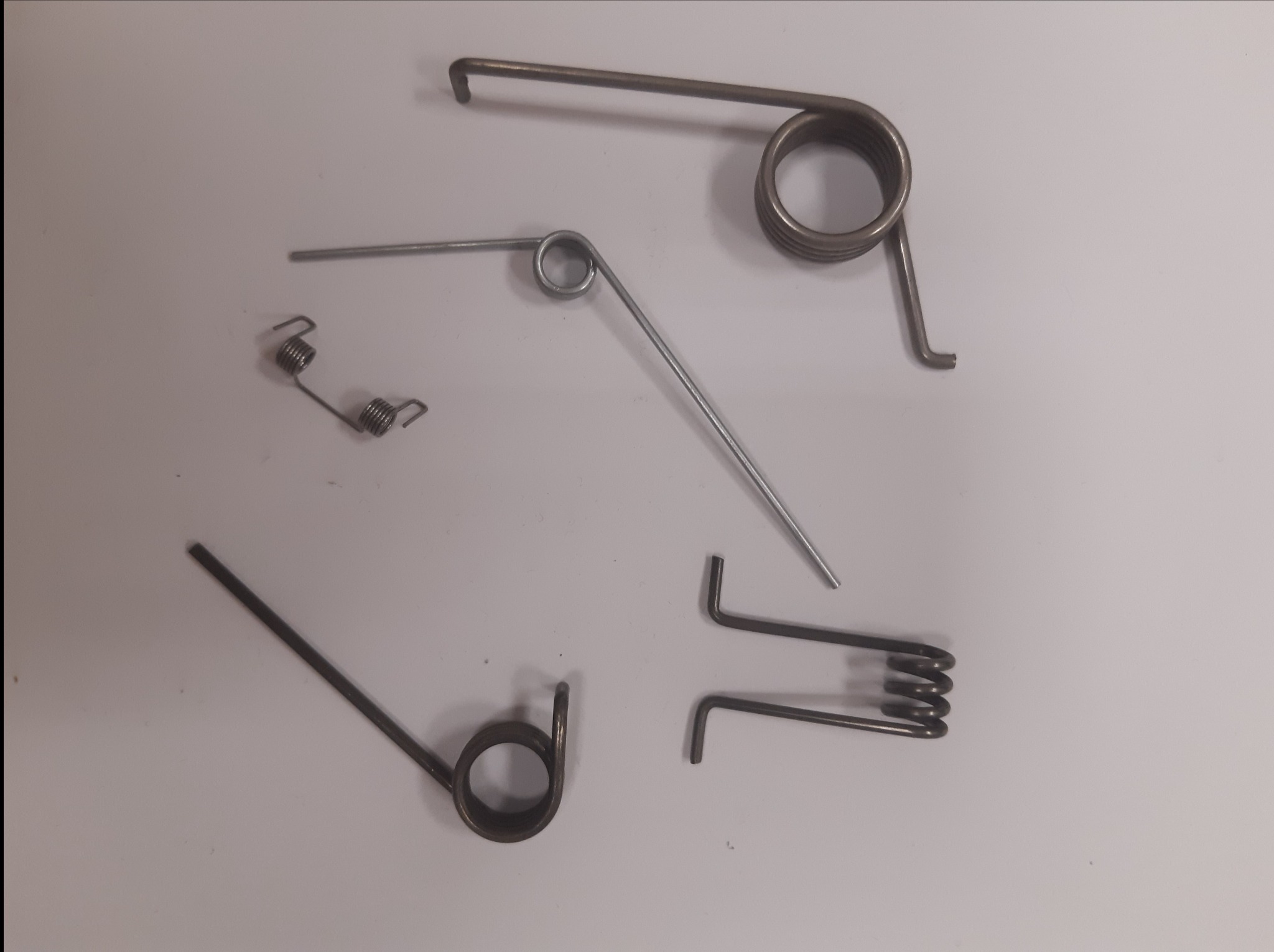 Zestaw różnorodnych sprężyn naciągowych i skrętnych, w tym spiralne i hakowe, ułożonych na jasnym tle. Widoczne detale konstrukcyjne i metaliczne wykończenie.