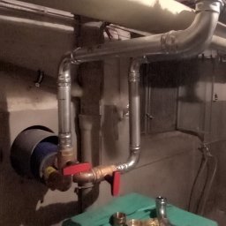 DN-INSTAL Natalia Mazurkiewicz - Instalacja rur z zaworami w piwnicy, widoczne złączki na zielonym materiale. Rury metalowe, sufit z widoczną izolacją. Ujęcie z dołu, podkreślające złożoność instalacji.