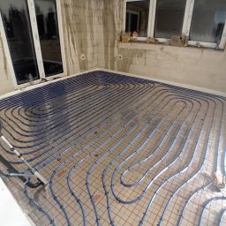 DN-INSTAL Natalia Mazurkiewicz - Niebieskie rury ogrzewania podłogowego ułożone na siatce w surowym wnętrzu z dwoma oknami. Widoczne narzędzia i materiały budowlane. Etap instalacji przed wylewką.