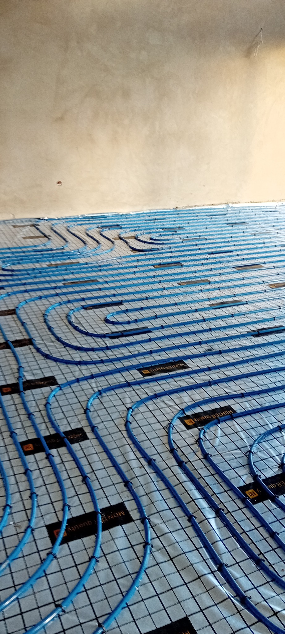 Niebieskie rury ogrzewania podłogowego ułożone na izolacji z siatką, widok z góry, gotowe do zalania posadzką. Układ spiralny rur.