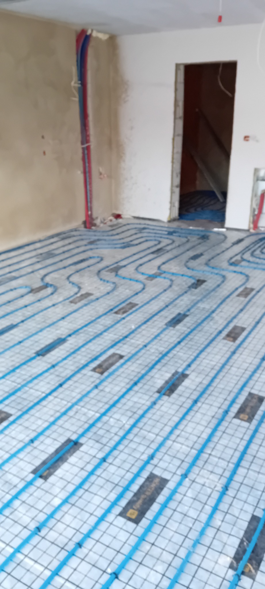 Niebieskie rury ogrzewania podłogowego ułożone na siatce w surowym wnętrzu. Widoczne pionowe rury przy ścianie i przygotowane przejście do innego pomieszczenia.