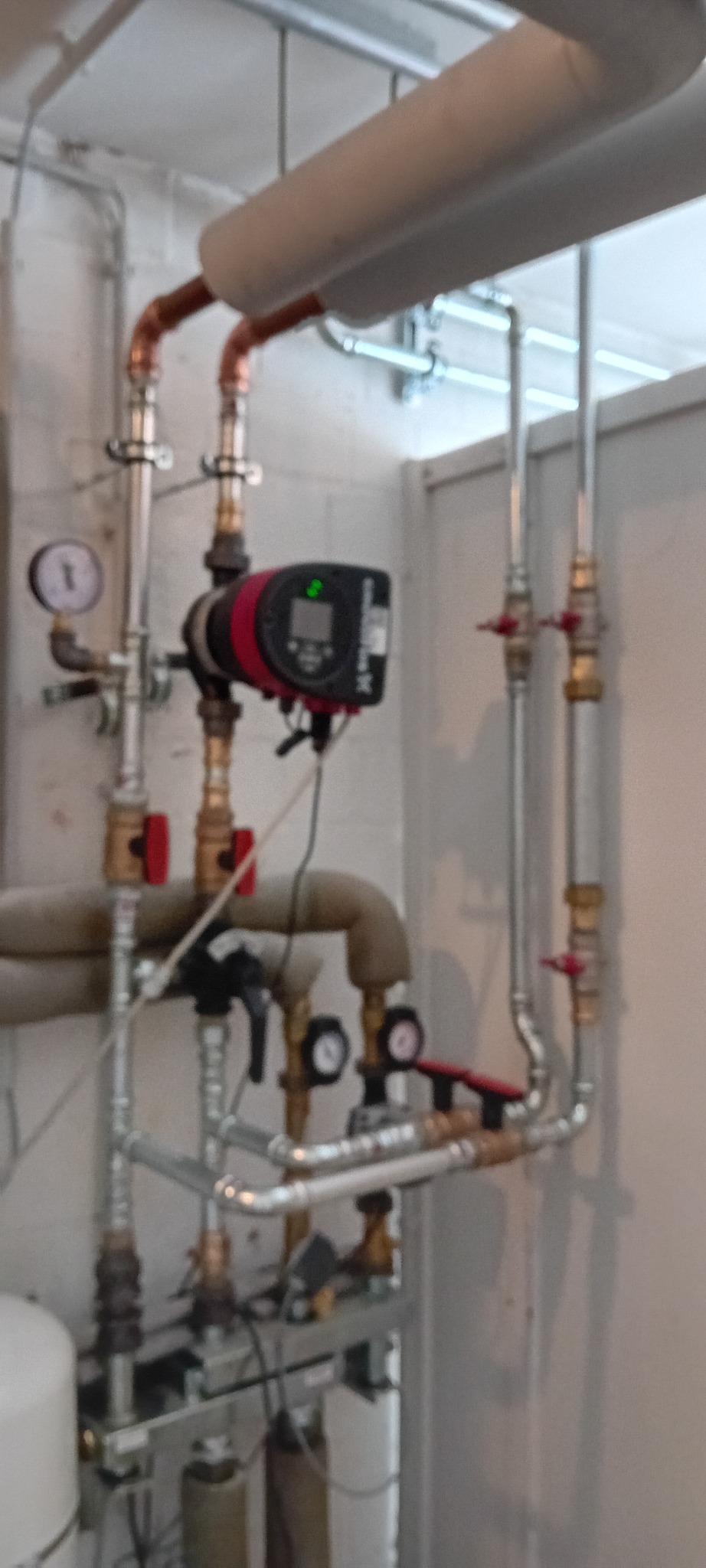 Złożona instalacja hydrauliczna z rur miedzianych i stalowych, zaworów, manometrów i pompy obiegowej w pomieszczeniu technicznym. Widoczne izolowane rury i elementy mocujące.