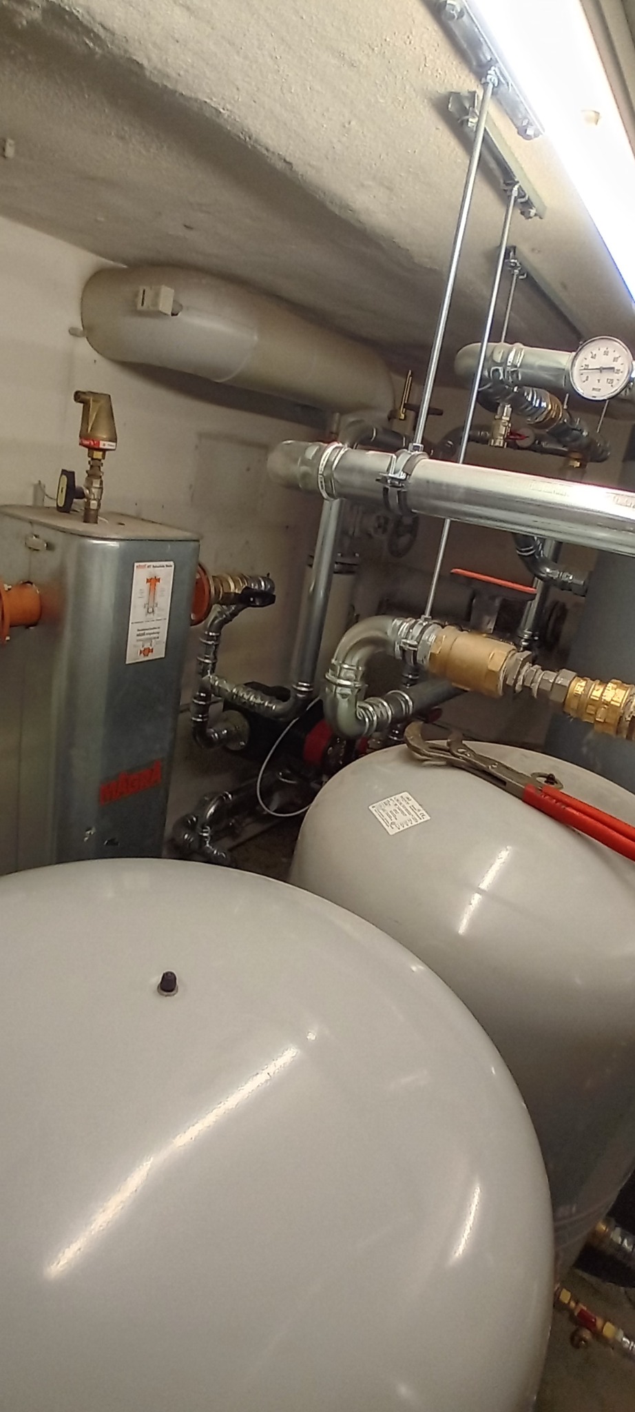 Złożona instalacja grzewcza z zbiornikami wyrównawczymi, rurami i manometrem. Widoczne narzędzia hydrauliczne. Elementy instalacji w piwnicy lub kotłowni.